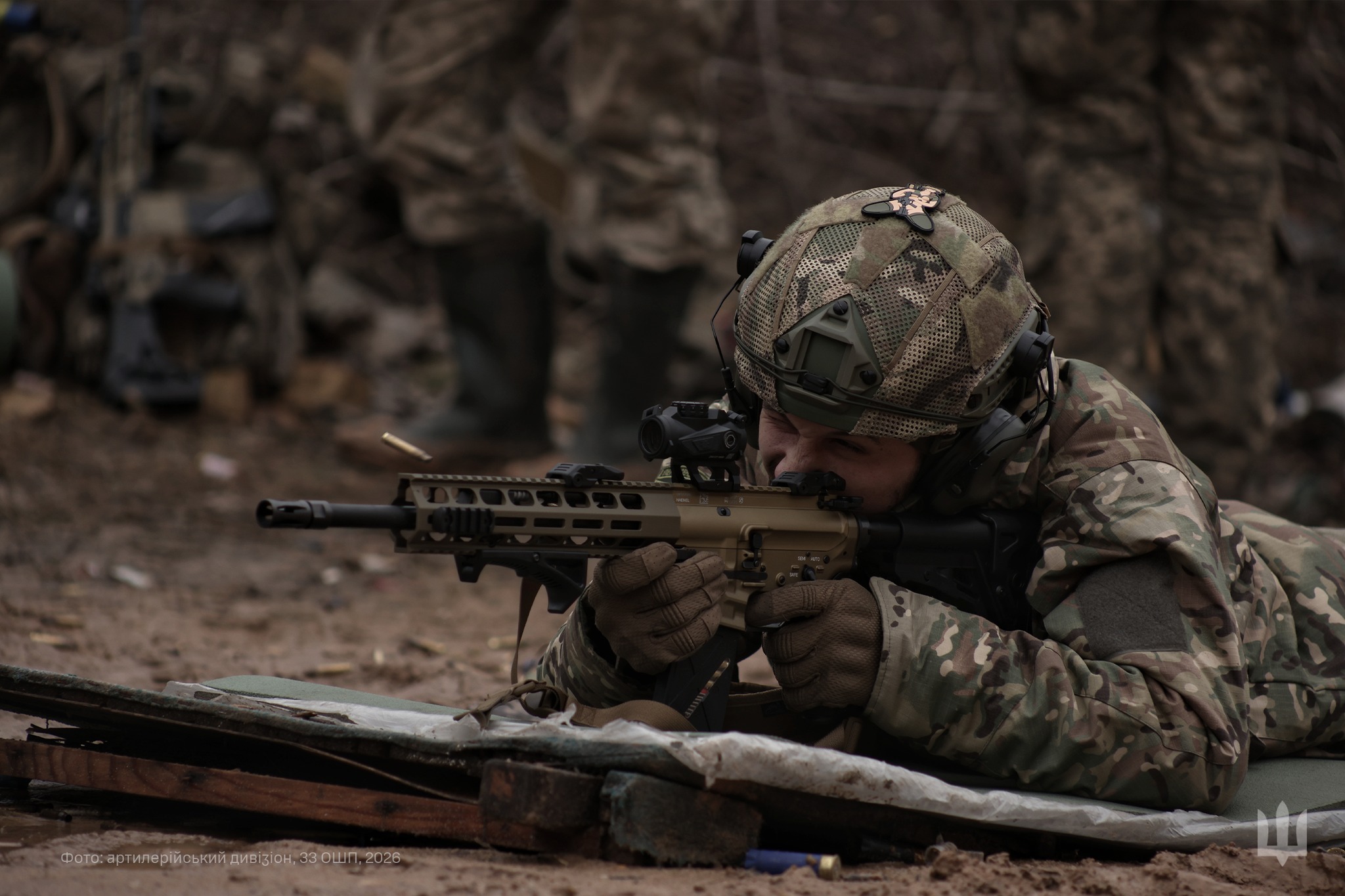 Український військовий у камуфляжі веде вогонь із автоматичної гвинтівки, лежачи на позиції за імпровізованим укриттям. На задньому плані видно інших бійців підрозділу, які перебувають на вогневій позиції.