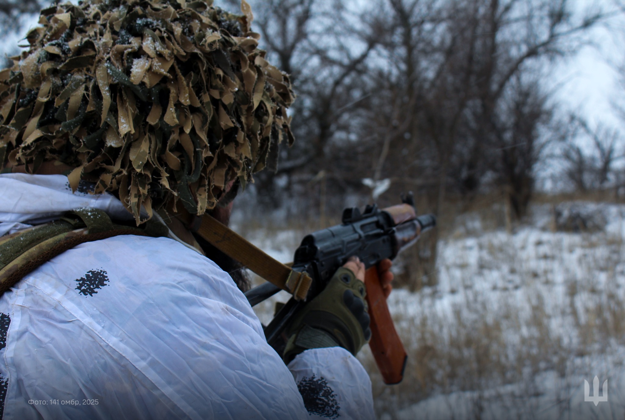 Боєць у зимовому маскувальному одязі та з камуфльованим шоломом веде вогонь з автомата у бік лісосмуги. Навколо видно засніжене поле та оголені дерева, що створюють суворий зимовий пейзаж на тлі бойових дій.