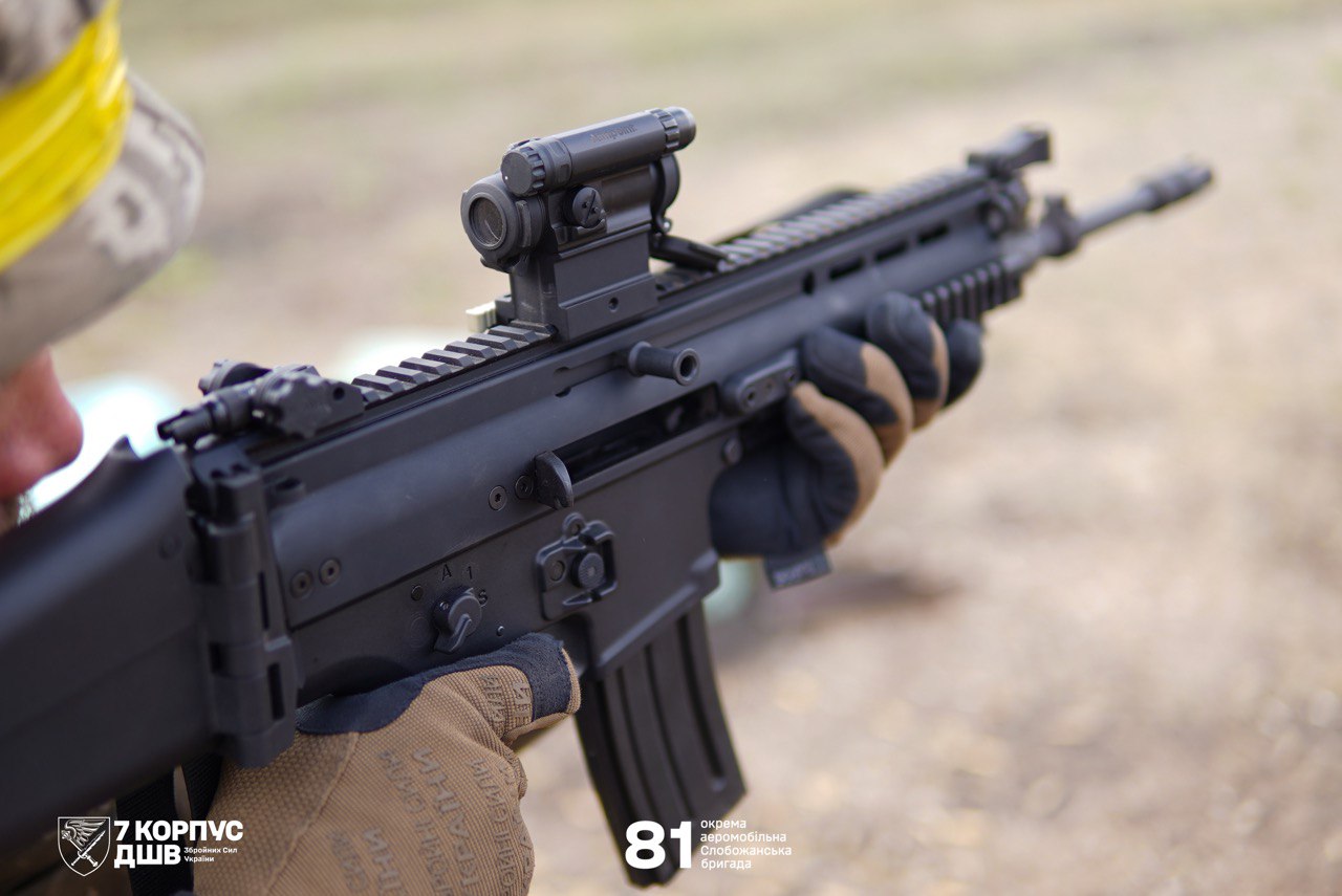 Крупним планом видно військового, який тримає сучасну штурмову гвинтівку з коліматорним прицілом. Боєць у рукавичках прицілюється, тримаючи зброю напоготові на відкритій місцевості.