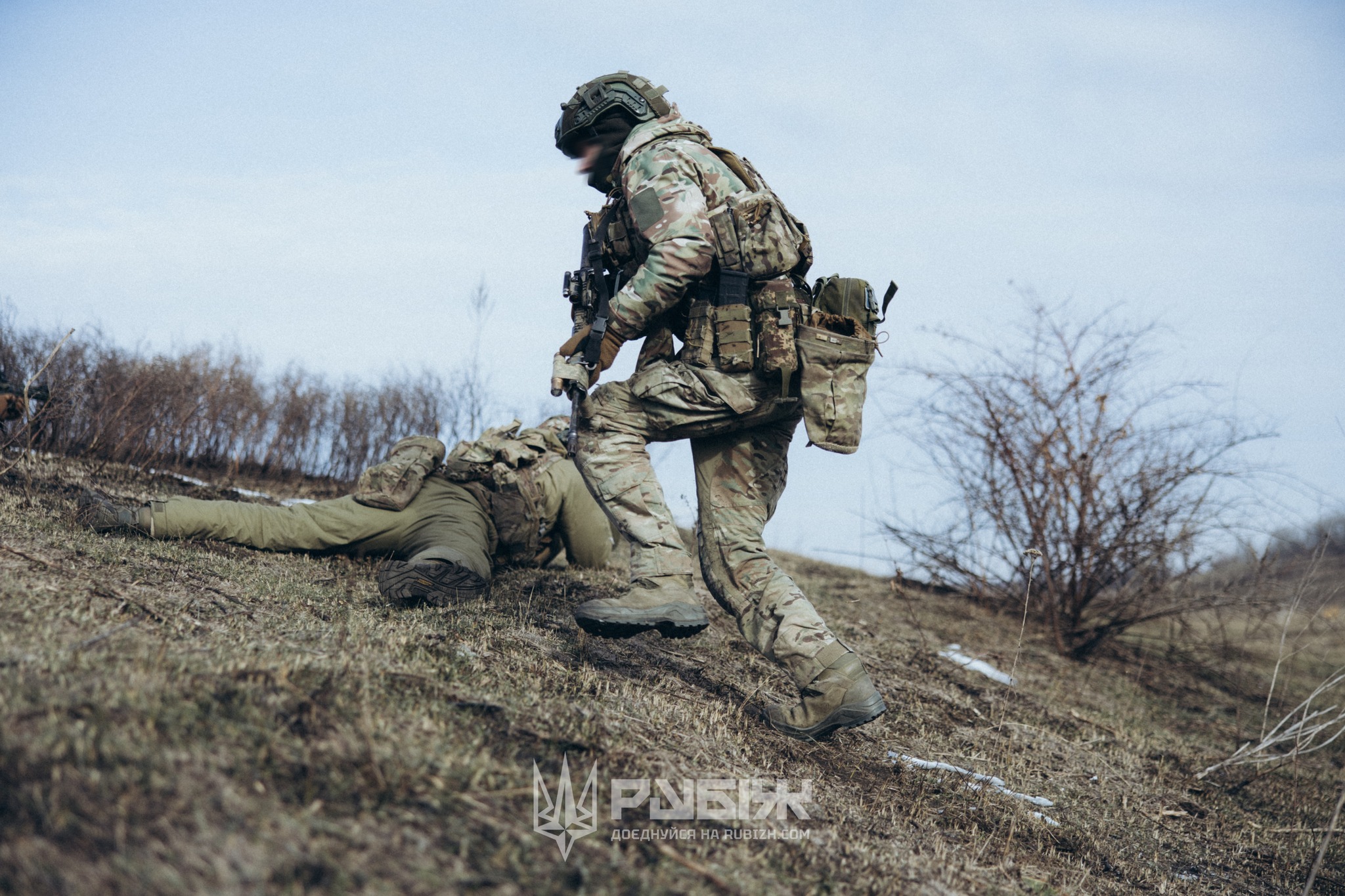 Боєць у повному спорядженні рухається схилом, прикриваючи побратима, який лежить на землі. Обстановка виглядає напруженою, а дії військового свідчать про виконання бойового або навчального завдання в польових умовах.