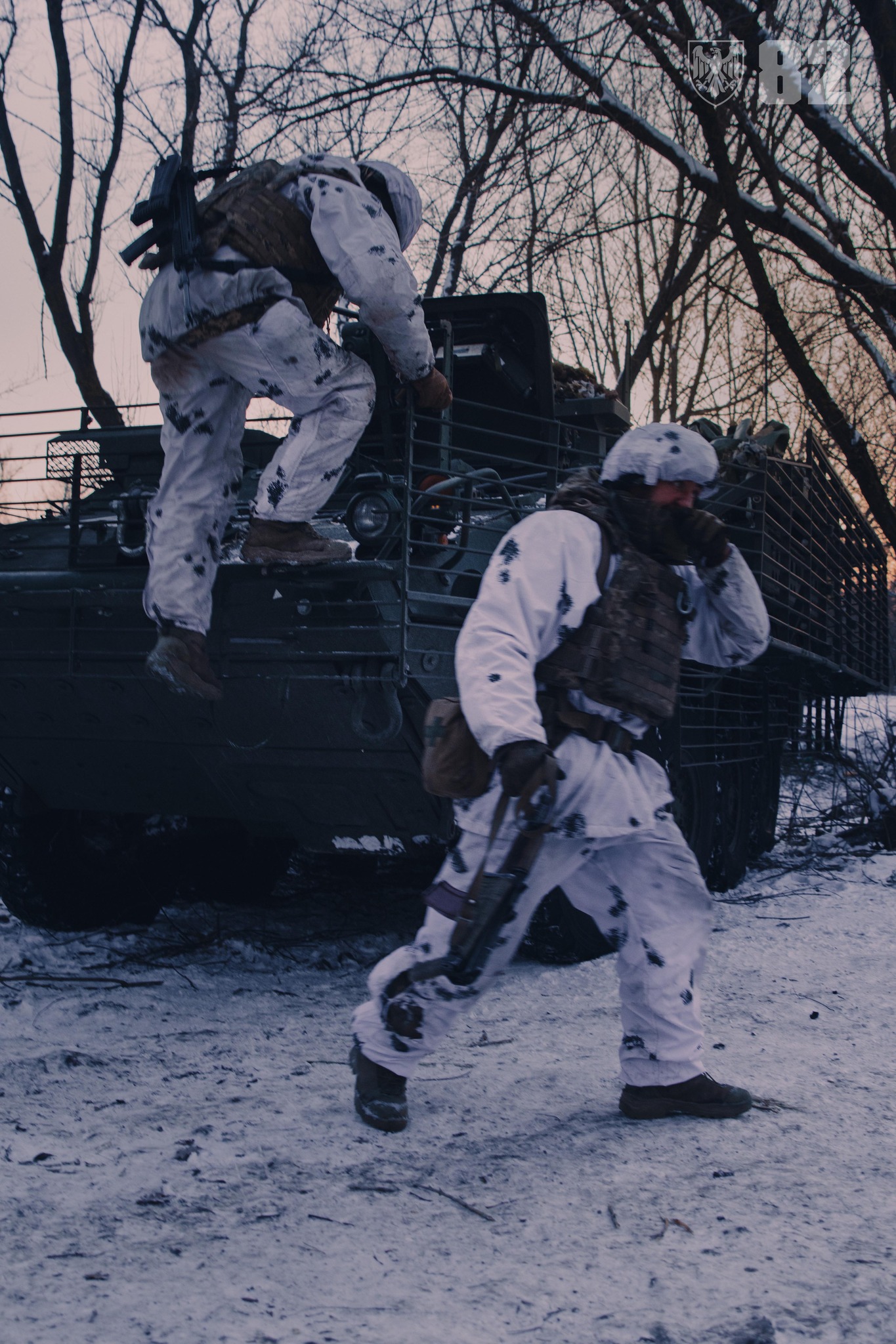 Двоє військових у зимовому маскувальному спорядженні залишають бронемашину на засніженій лісовій дорозі. Один боєць уже рухається вперед із автоматом у руках, тоді як інший спускається з техніки, готуючись приєднатися до руху.