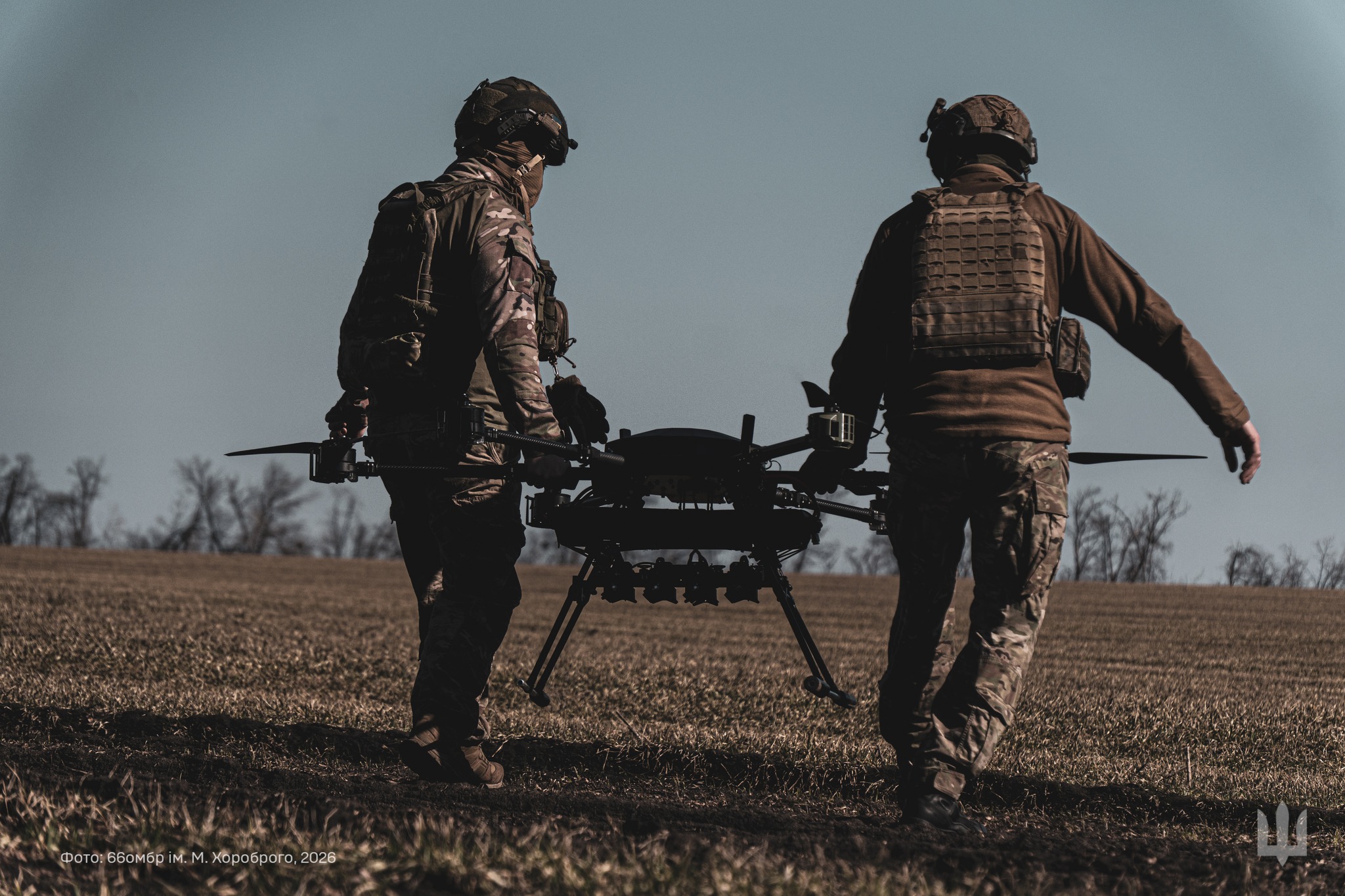 Двоє військових несуть великий безпілотник полем, готуючи його до розгортання. Їхнє спорядження та зосередженість свідчать про підготовку до виконання бойового завдання із застосуванням дрона.