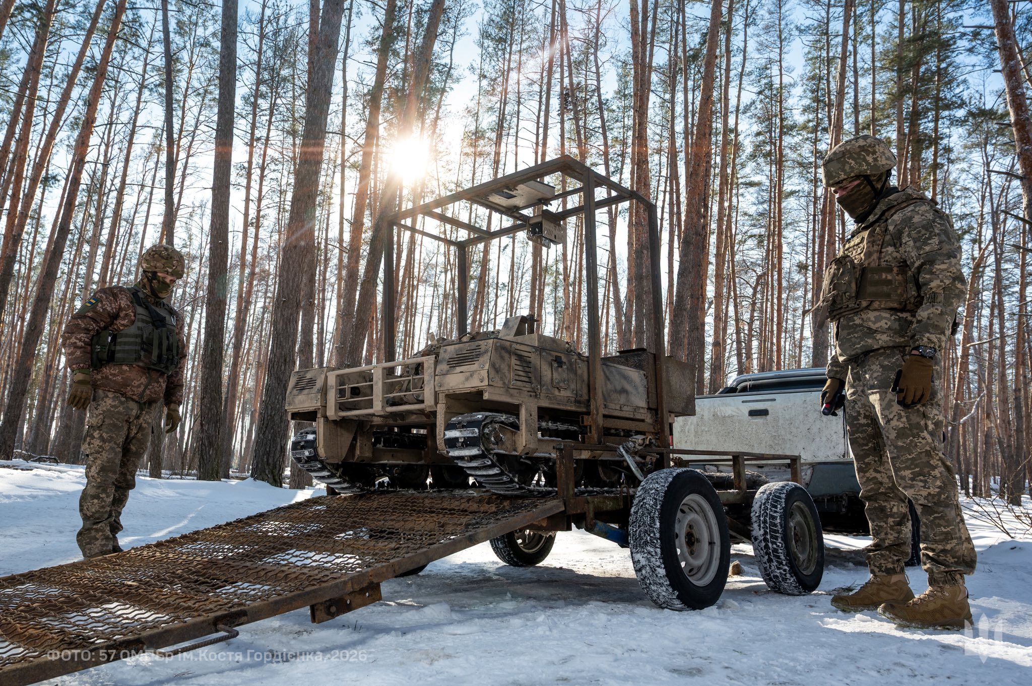 Двоє військових у зимовому лісі стоять поруч із причепом, на який завантажено гусеничний наземний робот. Сонце пробивається крізь дерева, підсвічуючи техніку та сніг, поки бійці контролюють процес розвантаження.