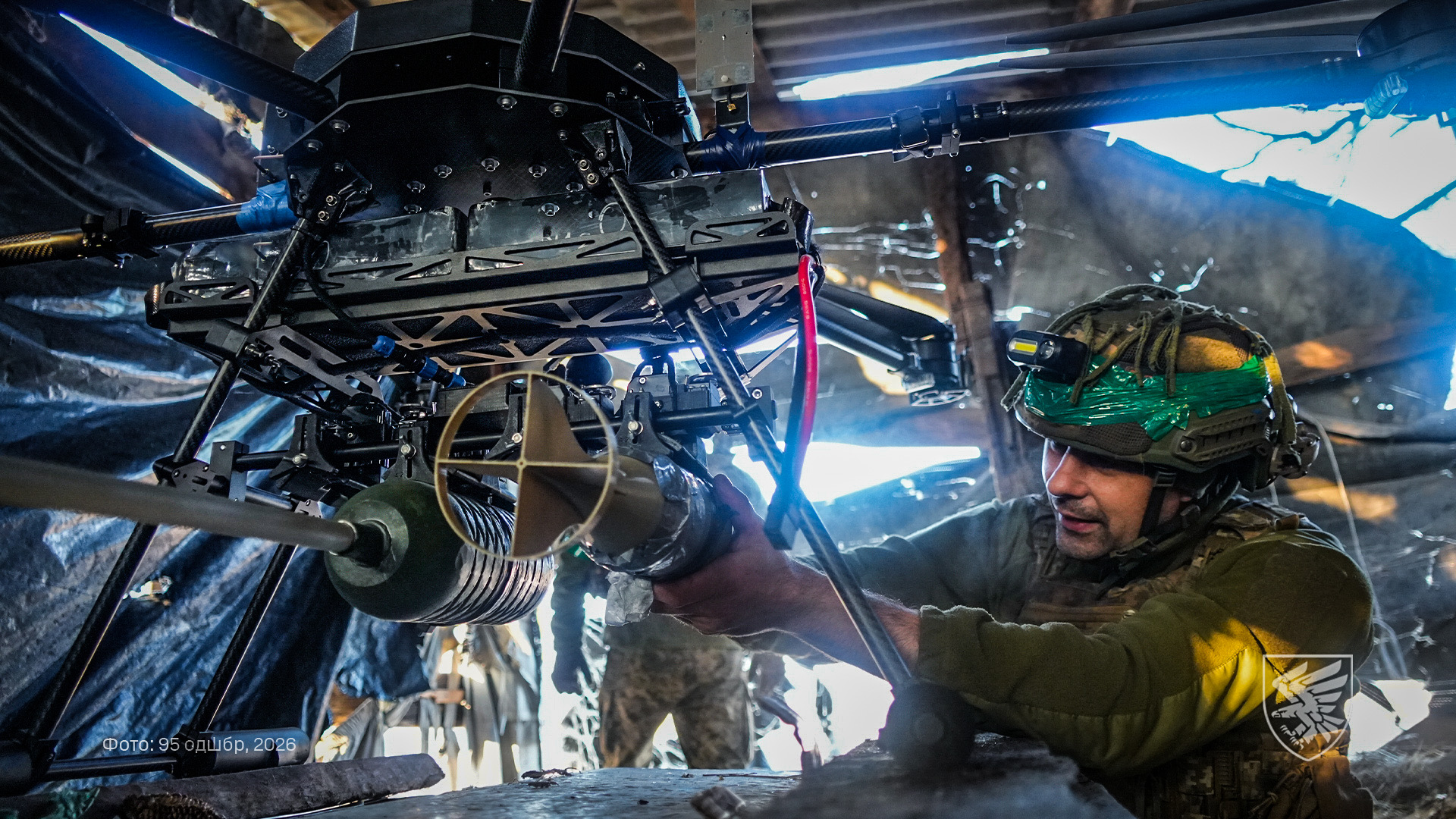Військовий у шоломі встановлює боєприпас на великий безпілотник у приміщенні, схожому на укриття. Дрон оснащений масивною конструкцією та підвісом, що свідчить про його призначення для важких навантажень.