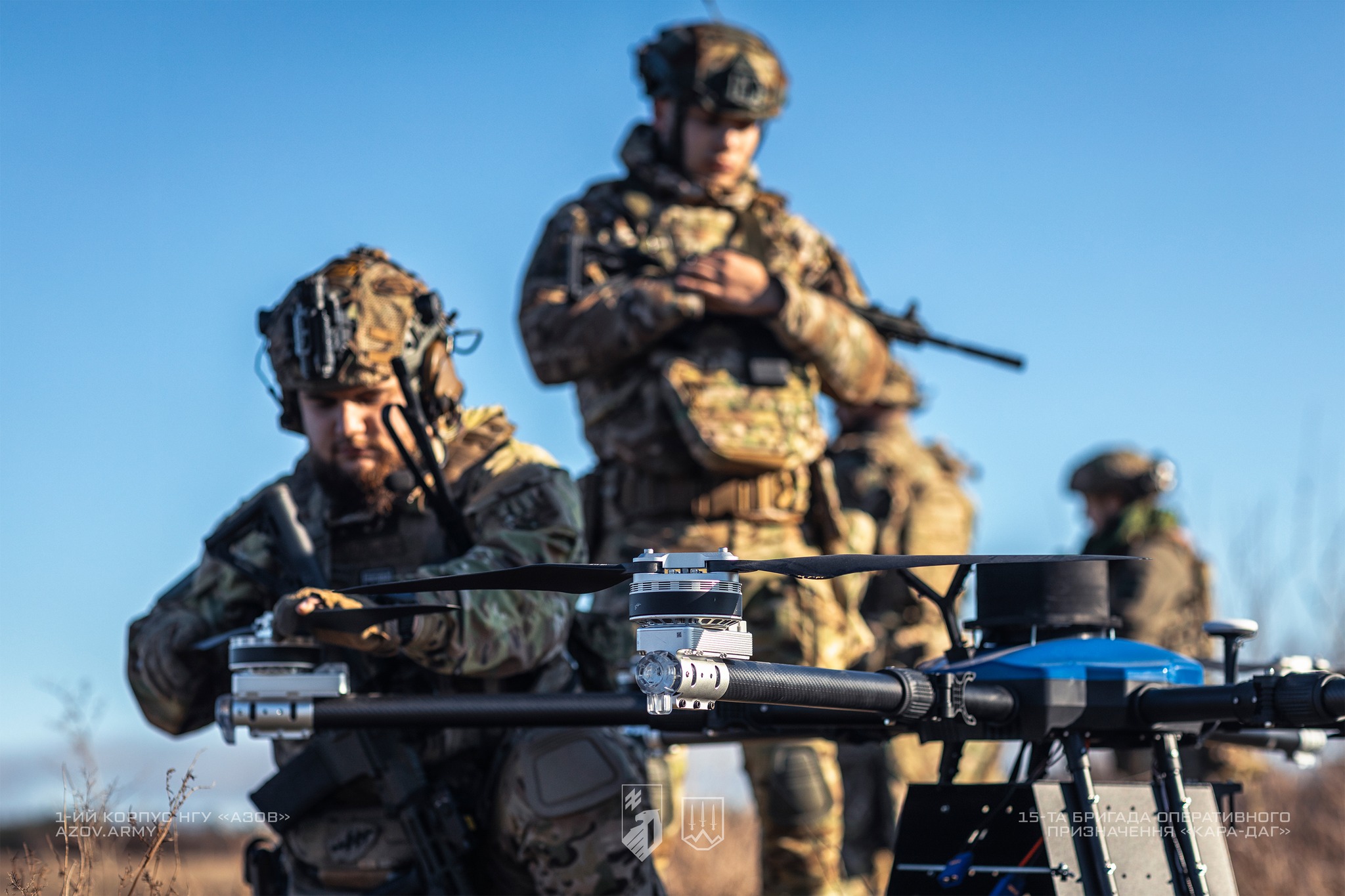 Українські військові у повному спорядженні готують до роботи великий дрон-бомбер, встановлений на землі перед ними. Один боєць налаштовує апарат, тоді як інші стоять поруч зі зброєю, контролюючи обстановку на відкритій місцевості.