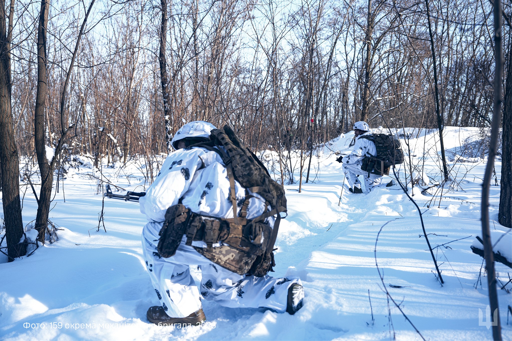 Двоє військовослужбовців у зимовому маскуванні пересуваються засніженим лісом, тримаючи зброю напоготові. Вони рухаються слідом один за одним, використовуючи рельєф і рослинність для прихованого просування.