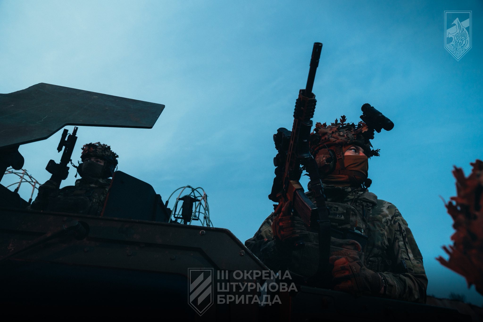 Двоє військових у повному спорядженні перебувають на бронетехніці, пильно оглядаючи місцевість і тримаючи зброю напоготові. Сутінкове небо створює напружену атмосферу, підкреслюючи їхню зосередженість і готовність до дій.