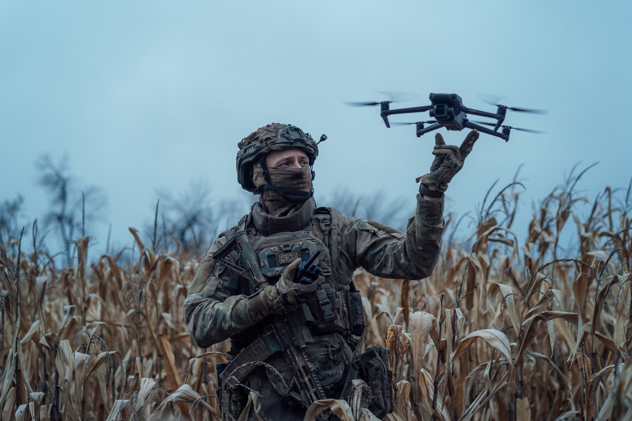 Військовий у повному спорядженні стоїть серед сухого кукурудзяного поля та керує невеликим квадрокоптером, який завис у повітрі перед ним. В одній руці він тримає пульт управління, а іншою спрямовує дрон, уважно стежачи за його польотом.