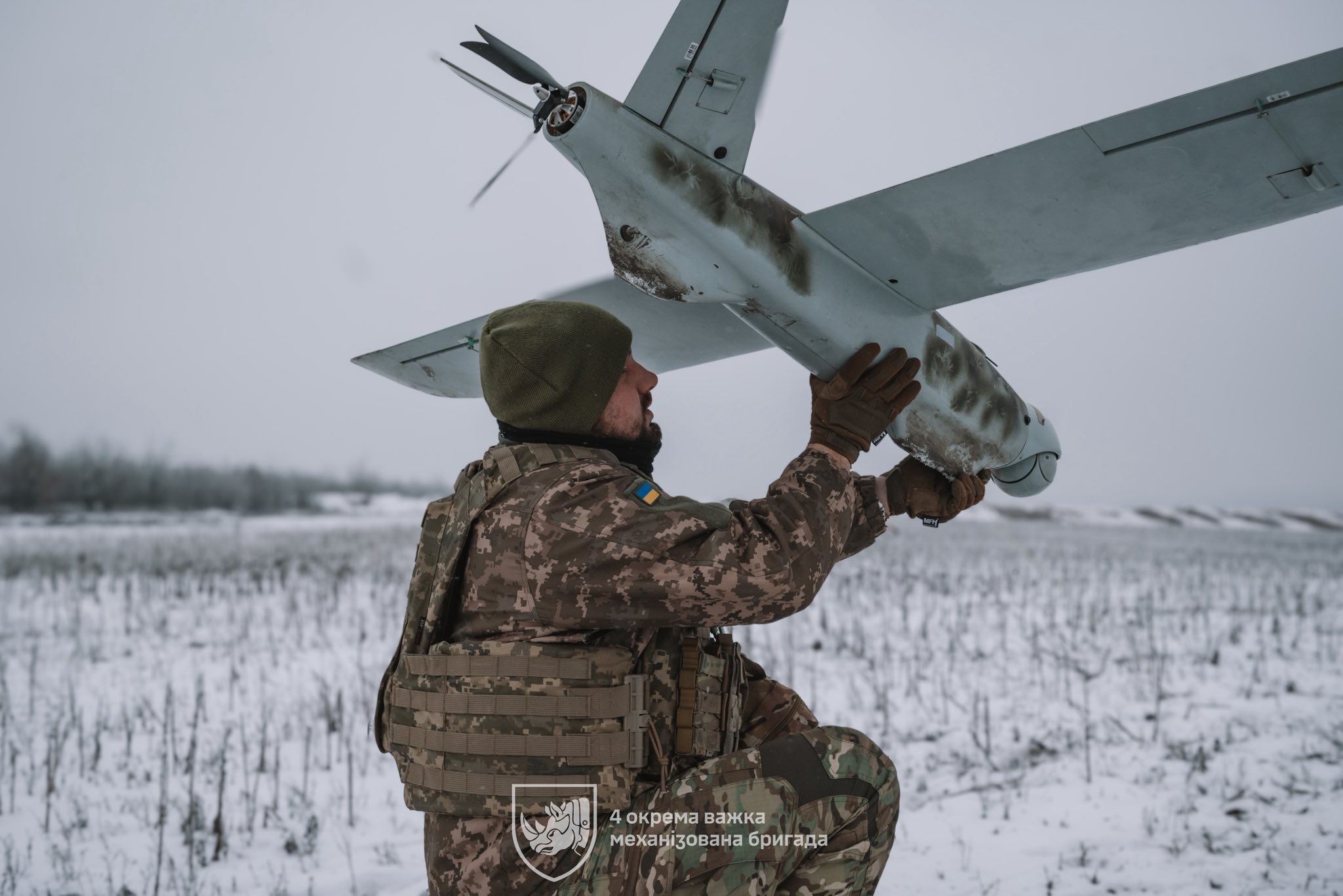 Український військовий у зимовому полі тримає в руках безпілотник літакового типу, готуючи його до запуску. На формі бійця видно шеврон із прапором України, а позаду простягається засніжений відкритий простір.