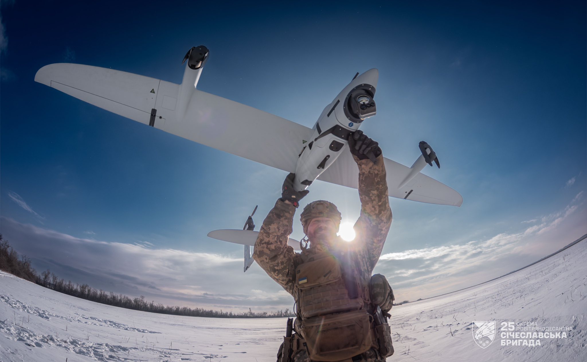 Військовослужбовець у зимовому полі тримає над головою розвідувальний безпілотник типу «крило», готуючи його до запуску. Сонце світить позаду бійця, підсвічуючи силует дрона на тлі ясного неба та засніженого ландшафту.