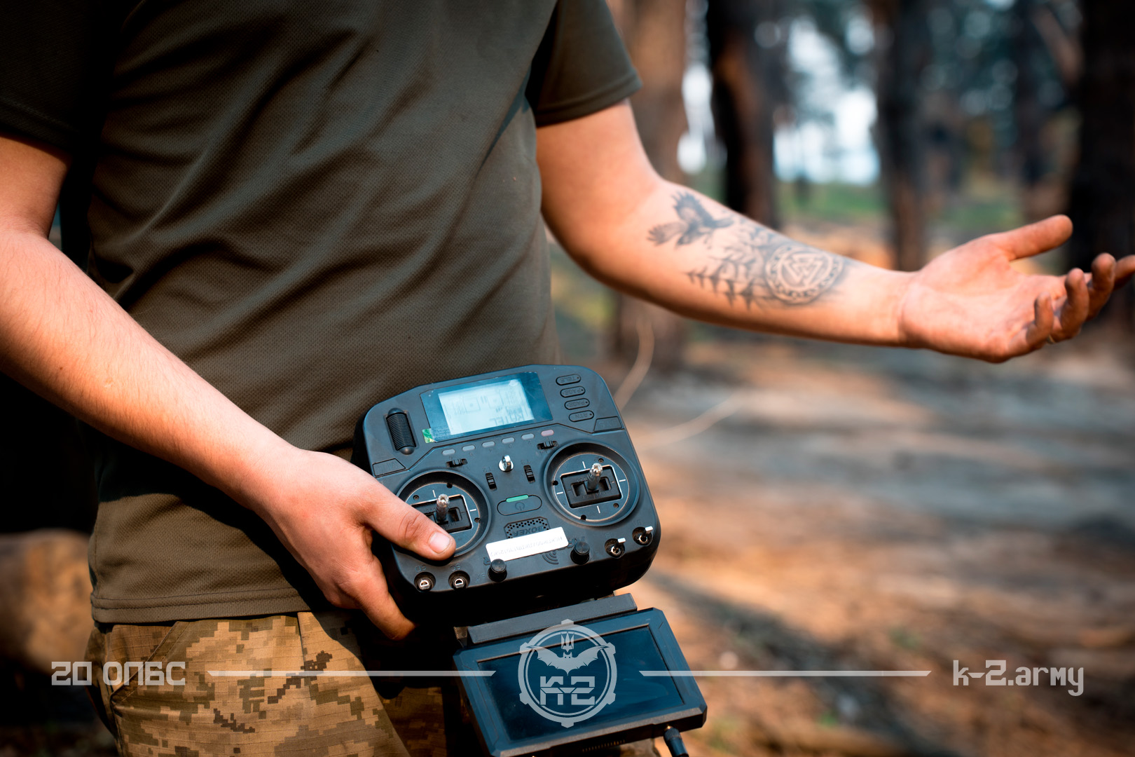 Військовослужбовець у польовій формі тримає в руках пульт керування безпілотником, стоячи серед лісу. На його передпліччі видно татуювання, а екран апаратури відкритий для контролю польоту дрона.