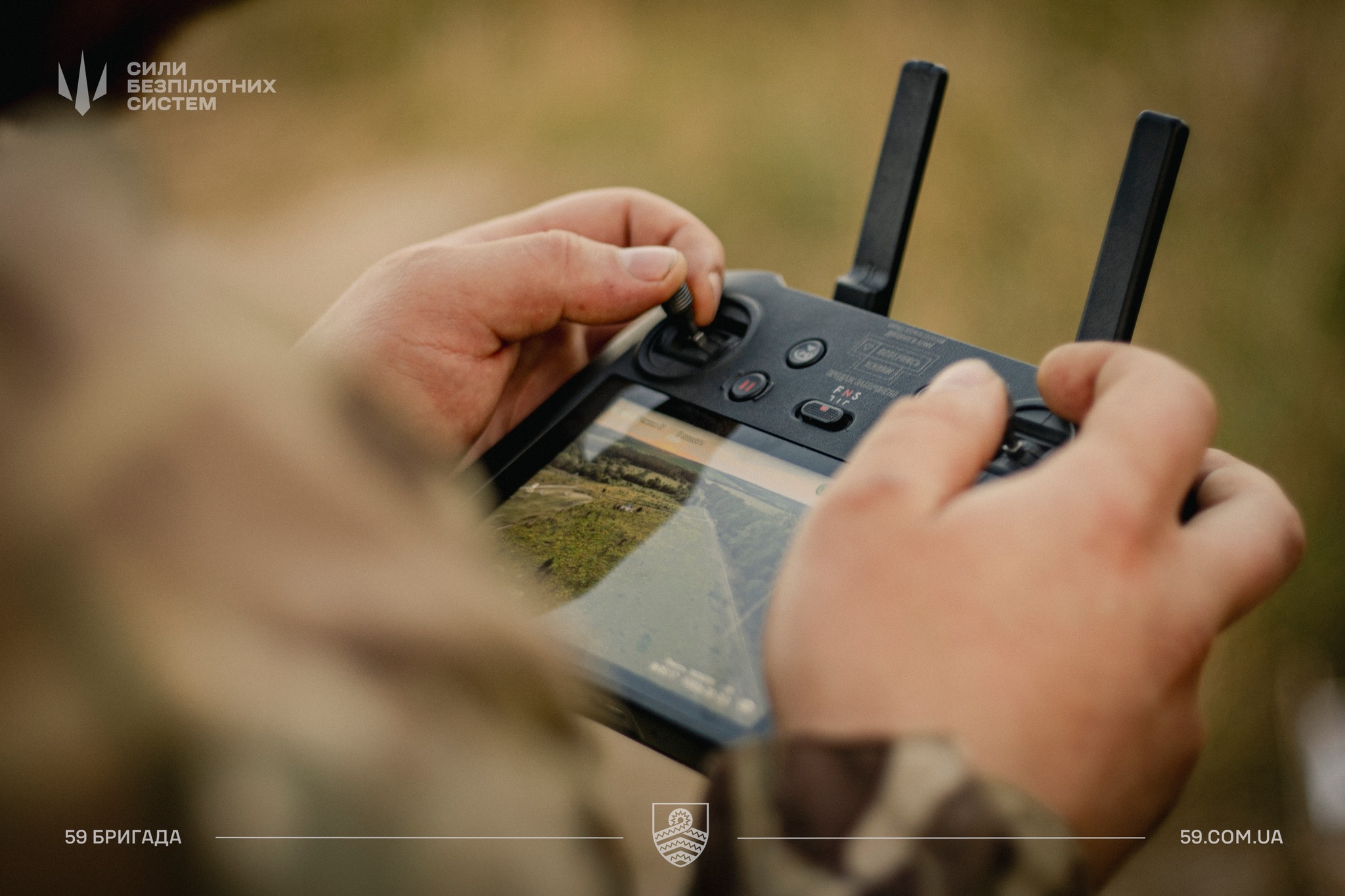 Крупним планом видно руки військового, який тримає пульт керування безпілотником із двома антенами. На екрані контролера відображається зображення місцевості з камери дрона під час польоту.