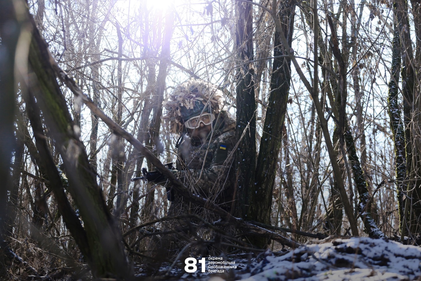 Український військовий у камуфляжі та захисних окулярах займає позицію серед густих дерев і гілок у зимовому лісі. Крізь гілля пробивається сонячне світло, підсвічуючи бійця, який уважно стежить за місцевістю зі зброєю напоготові.