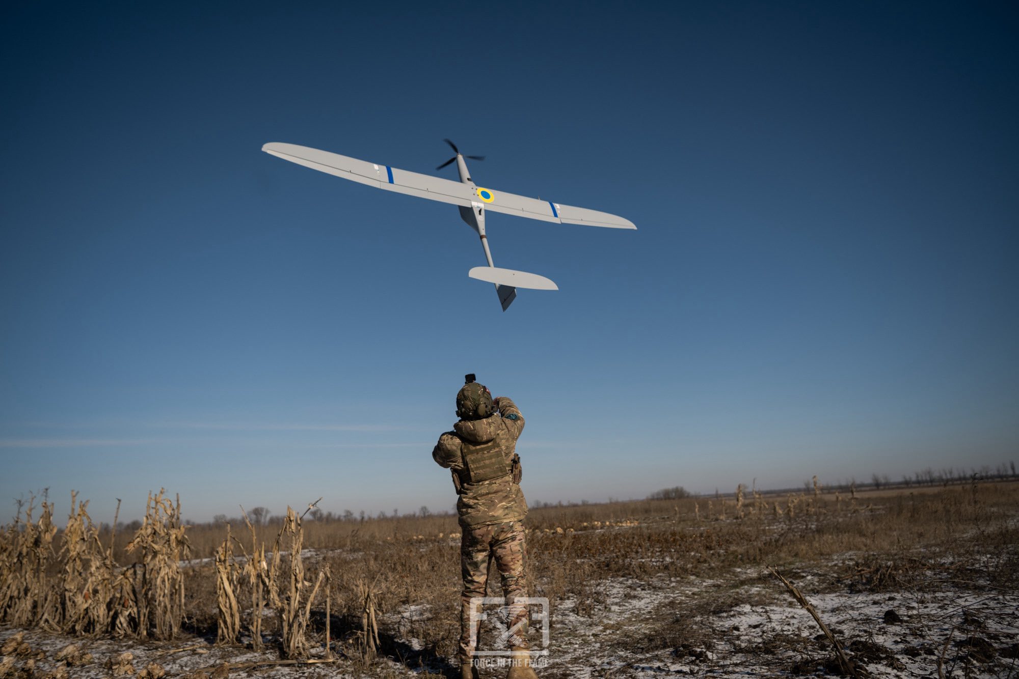 Військовослужбовець у польових умовах запускає безпілотник літакового типу, спрямовуючи його в небо над відкритою місцевістю. Апарат із українськими розпізнавальними знаками набирає висоту над засніженим полем і сухою рослинністю.