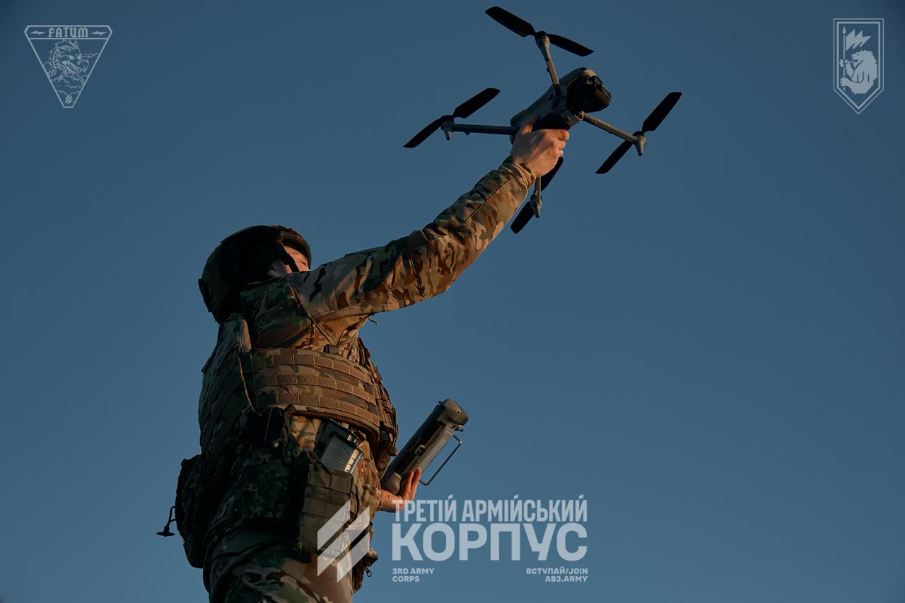 Військовий у спорядженні піднімає квадрокоптер над головою, готуючись до його запуску в небо. Чистий фон і чіткий силует підкреслюють момент початку роботи безпілотника.