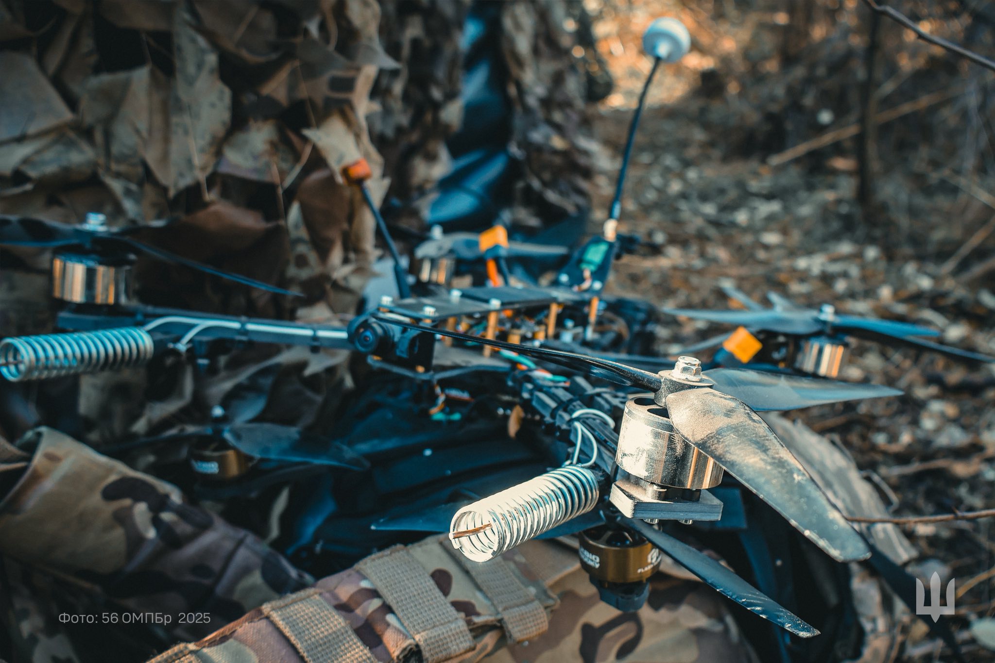 На передньому плані видно бойовий квадрокоптер із встановленими антенами та підсилювачами сигналу, підготовлений до роботи в польових умовах. Поруч перебувають військові в камуфляжі, які, ймовірно, налаштовують або обслуговують дрон у лісистій місцевості.