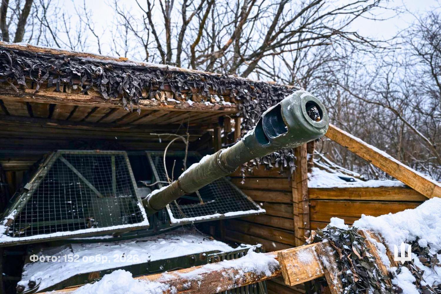 Зі снігового укриття, зробленого з дерев’яних балок і замаскованого сіткою, виступає довгий ствол бронетехніки, спрямований у бік лісосмуги. Машина стоїть під навісом серед зимового пейзажу, де голі дерева й шар снігу підкреслюють польові умови позиції.