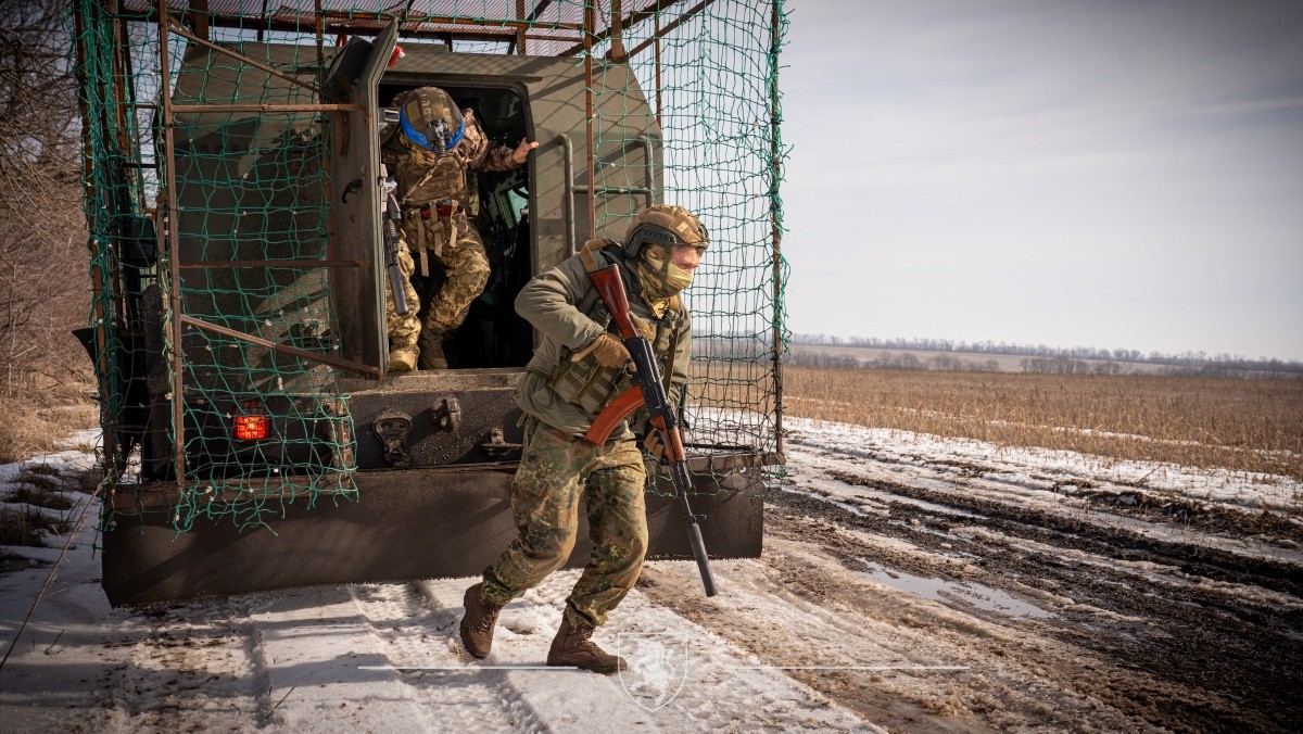 Військові висаджуються з бронемашини на засніженій польовій дорозі, швидко займаючи позиції. Один із бійців із автоматом прикриває рух, тоді як інший виходить із транспорту, готуючись приєднатися до дій.