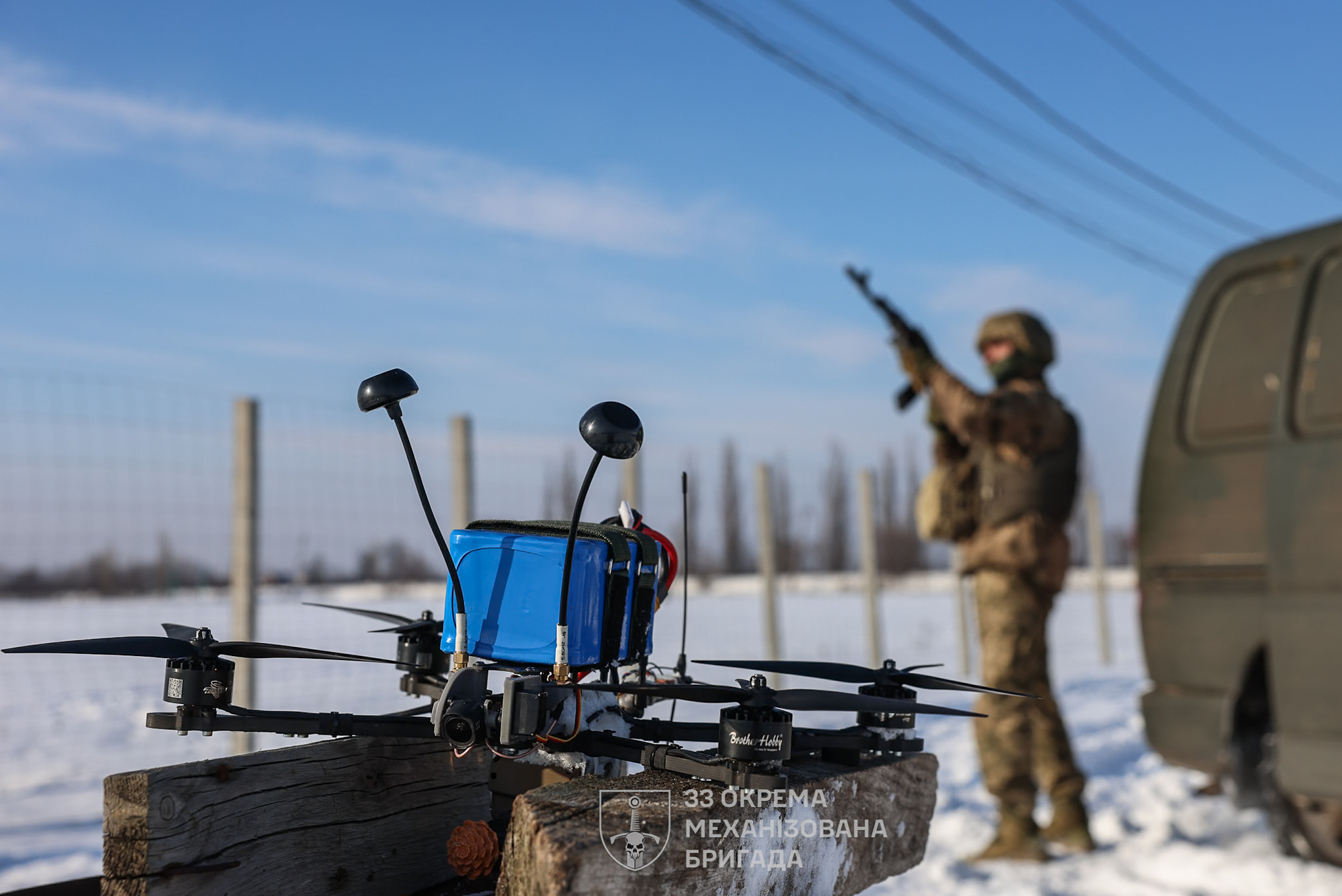 На передньому плані стоїть квадрокоптер із антеною та акумулятором, підготовлений до запуску в польових умовах. На задньому плані, розмитий, військовий із автоматом перебуває поруч із автомобілем на засніженій місцевості.