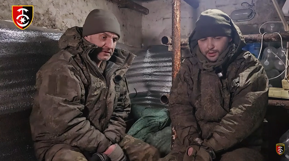 Двоє чоловіків у брудному військовому одязі сидять у тісному укритті або бліндажі з металевими стінами. На їхньому одязі закріплені мікрофони, що свідчить про запис інтерв’ю або відеосвідчень.