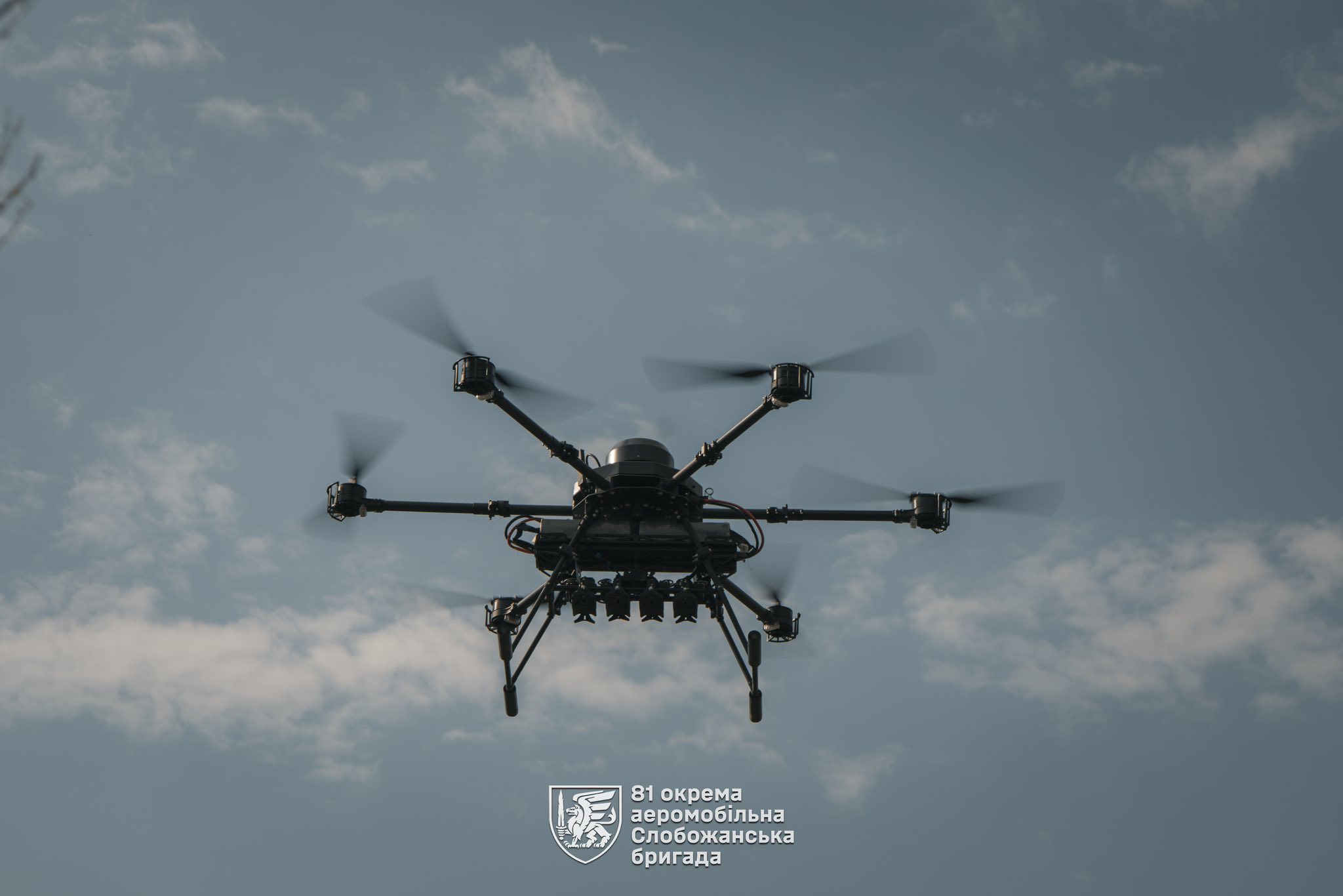 У небі летить багатороторний безпілотник із кількома гвинтами, який утримується в повітрі на тлі легких хмар. Під корпусом дрона видно підвішену апаратуру, ймовірно призначену для виконання бойових або розвідувальних завдань.