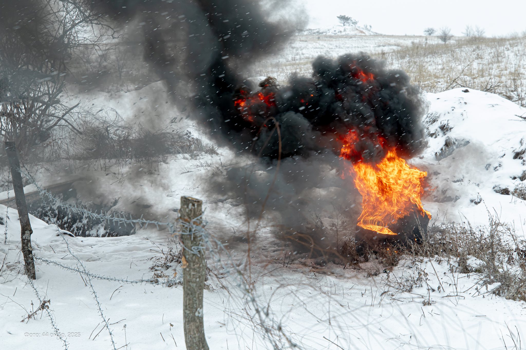 На засніженій місцевості палає підбита техніка, з якої виривається яскраве полум’я та густі клуби чорного диму. Вогонь і дим різко контрастують із білим зимовим пейзажем, а поряд видно колючий дріт і сліди боїв.