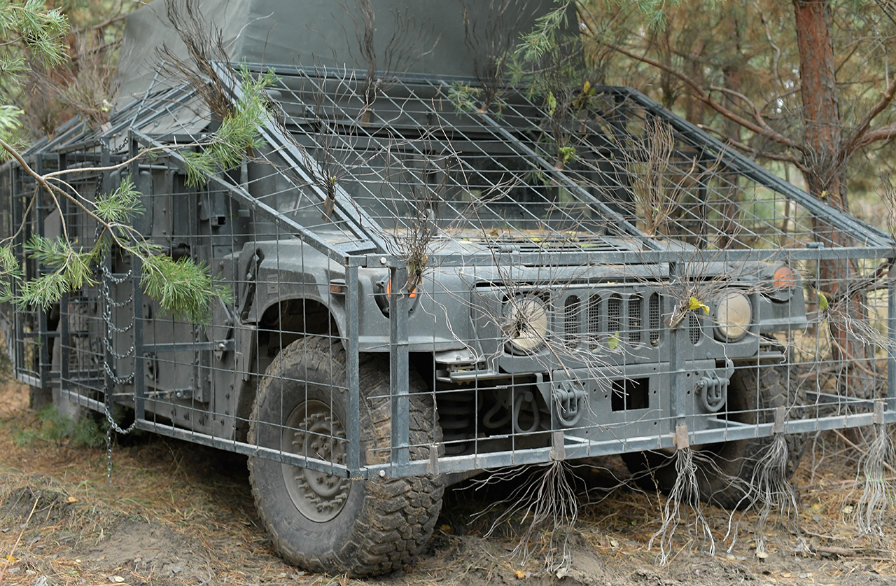 Військовий автомобіль стоїть у лісі, прихований серед сосен і гілля. Машина обладнана металевим захисним каркасом і маскувальними елементами для захисту від дронів та уламків.