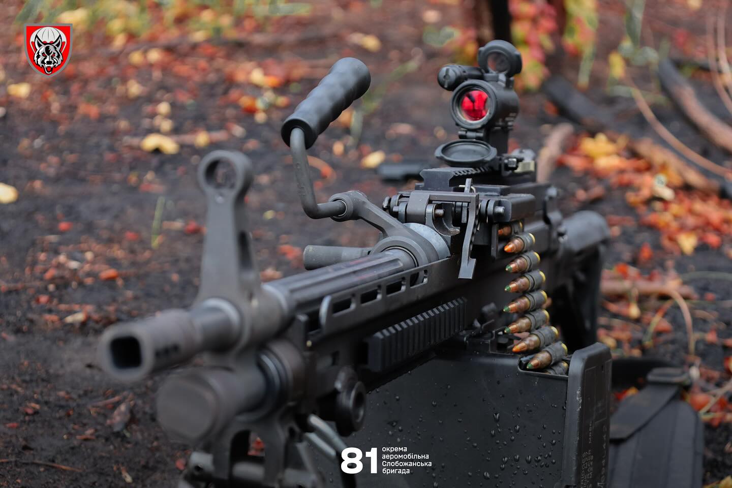 На передньому плані видно кулемет із оптичним прицілом, встановлений на землі серед осіннього листя. До зброї приєднана стрічка з набоями, що подається з коробки для боєприпасів.