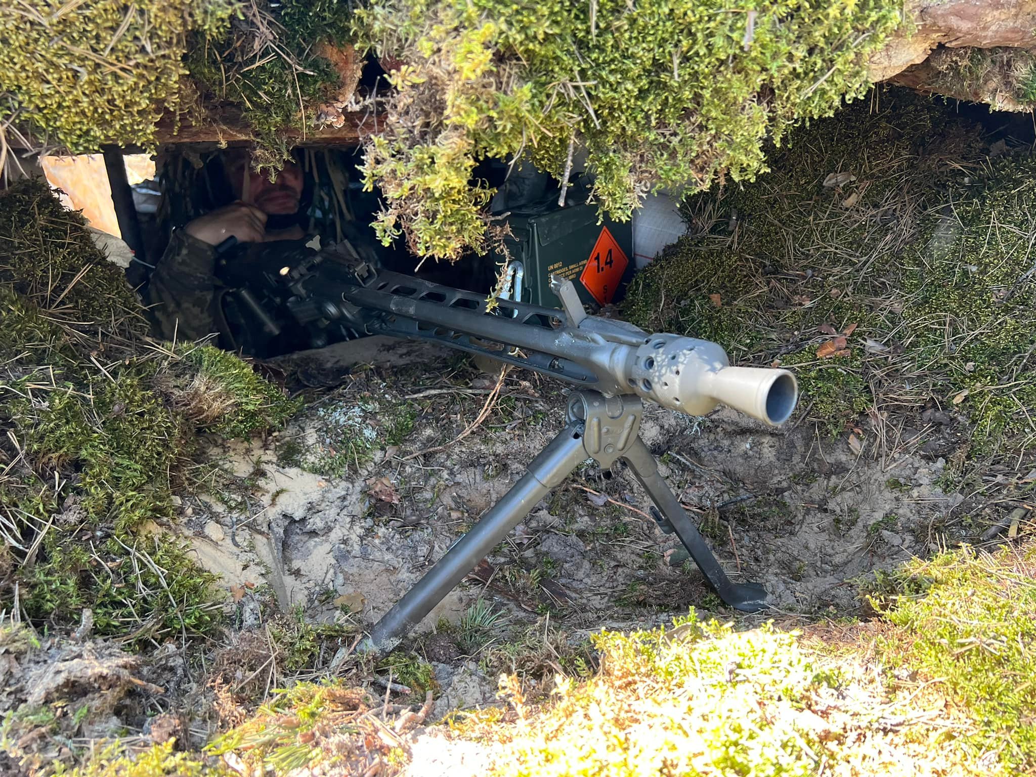 Військовий перебуває у замаскованому укритті, обладнаному в землі та прикритому дерном і гілками. Перед входом встановлено кулемет на тринозі, спрямований у бік можливого противника.