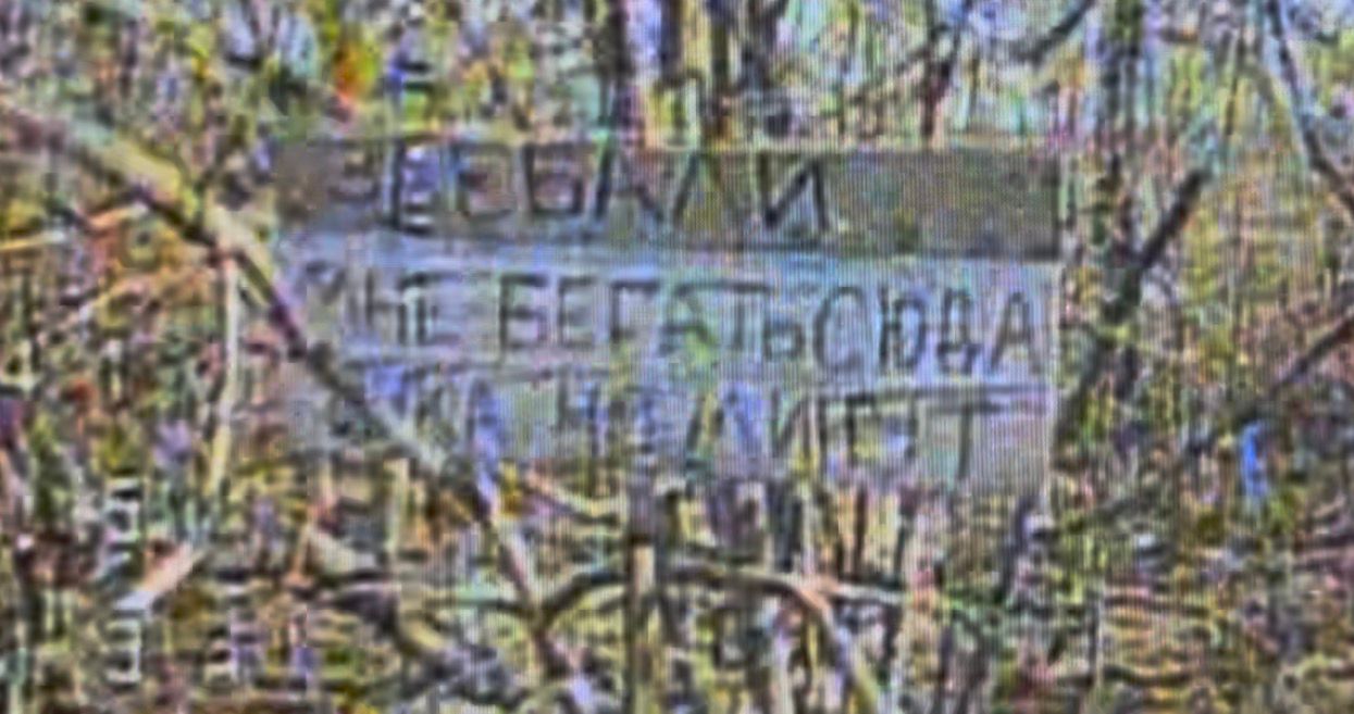 На зображенні видно картонний аркуш із написом, розміщений серед гілок і рослинності. Текст частково розмитий, але виглядає як імпровізований напис або позначка, залишена в польових умовах.