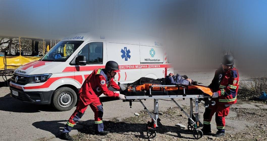 Медики у червоній формі перевозять пораненого на ношах до карети швидкої допомоги з написом «Екстрена медична допомога». Автомобіль стоїть поруч на відкритій місцевості, а частина зображення навмисно розмита.