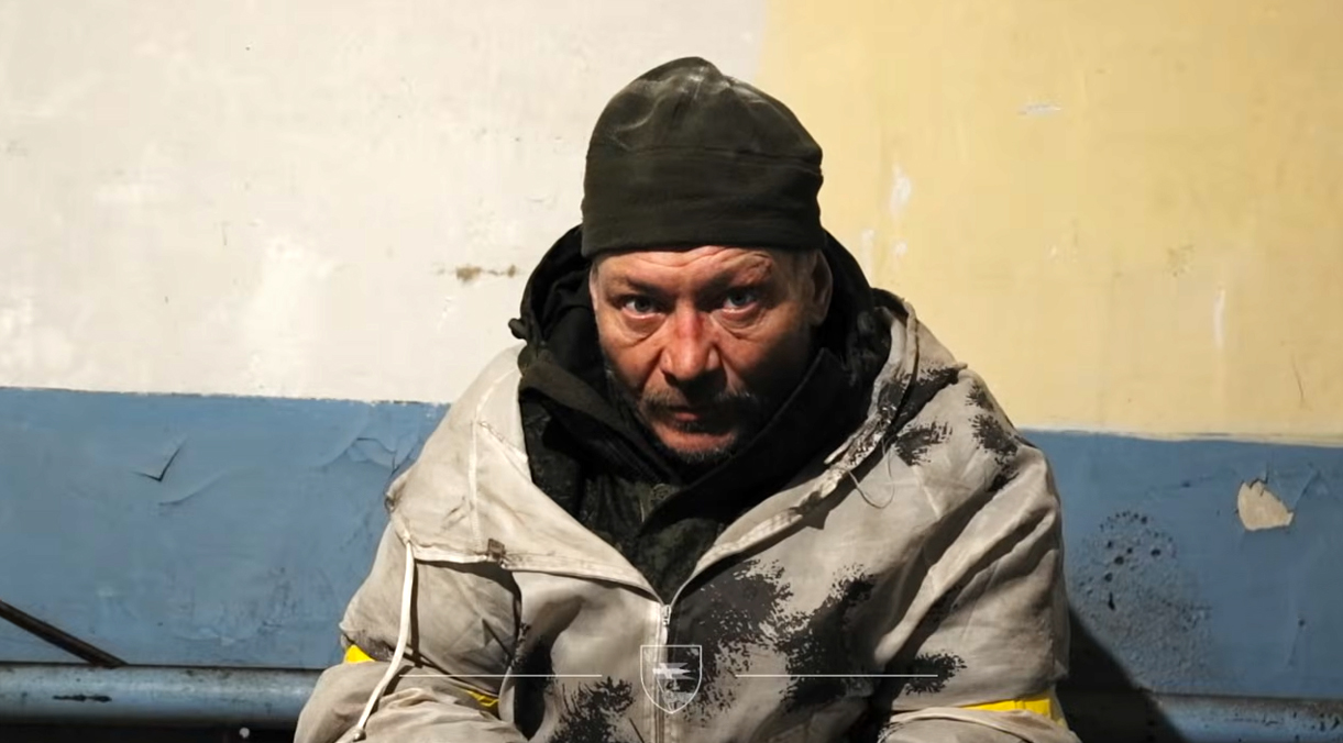 Чоловік у брудному військовому одязі та чорній шапці сидить у приміщенні з облупленими стінами, схожому на підвальне укриття або технічну кімнату. Його обличчя напружене й виснажене, а на рукаві видно жовту стрічку.