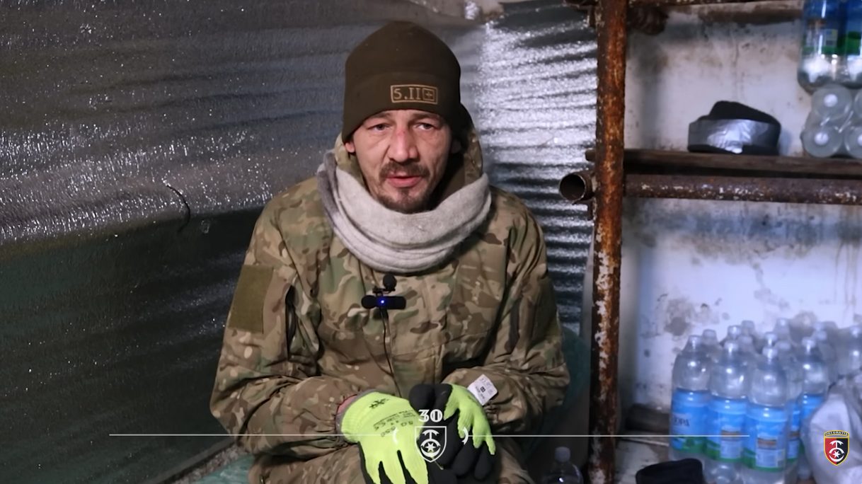 Чоловік у військовій формі сидить у приміщенні, схожому на укриття або бліндаж із металевими стінами. Поруч на полиці стоять кілька пляшок води, а на його одязі видно мікрофон, що свідчить про запис інтерв’ю або відеозвернення.