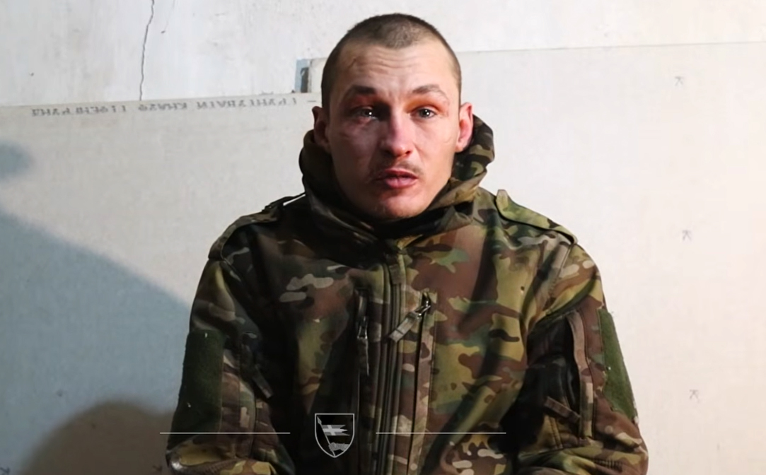 Чоловік у камуфляжній куртці сидить біля світлої стіни під час запису відео або інтерв’ю. Його обличчя виглядає втомленим і напруженим, а погляд спрямований трохи вбік від камери.