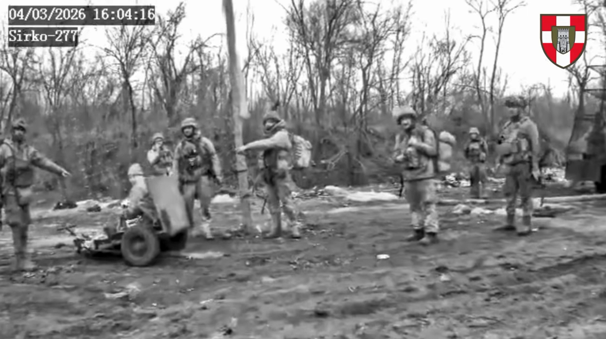 Група військових перебуває на відкритій місцевості серед зруйнованих дерев і багнюки. Один із бійців сидить на невеликому візку, тоді як інші військові навколо жестами щось пояснюють або координують дії.