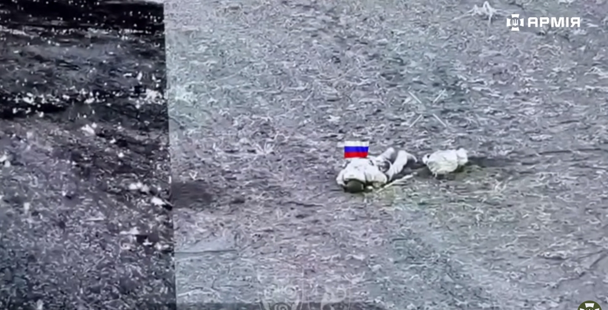 З висоти видно відкриту ділянку місцевості з нерівним рельєфом, де на землі лежить військовий поруч із розкиданим спорядженням. Поруч нанесено графічні позначки та символи, характерні для відеозйомки з безпілотника, що вказує на ведення спостереження або коригування.