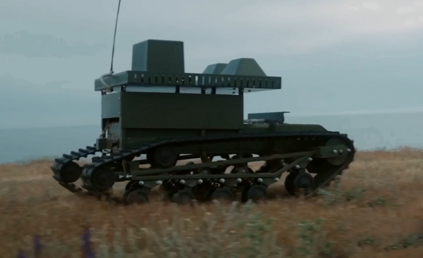 Гусеничний наземний роботизований комплекс рухається полем серед високої трави. На платформі встановлений броньований модуль із антеною, що свідчить про дистанційне керування машиною.