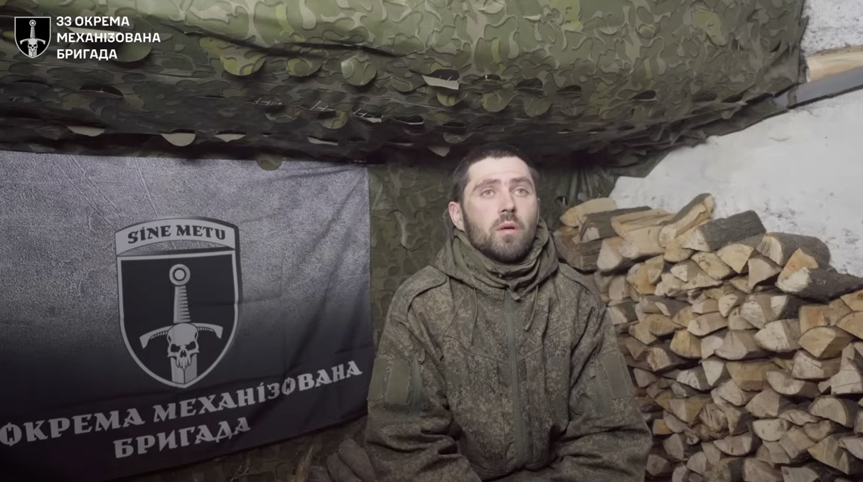 У невеликому укритті під маскувальною сіткою сидить чоловік у військовому одязі, поруч із ним складені дрова. Позаду видно прапор із символікою 33-ї окремої механізованої бригади та написом «Sine metu».