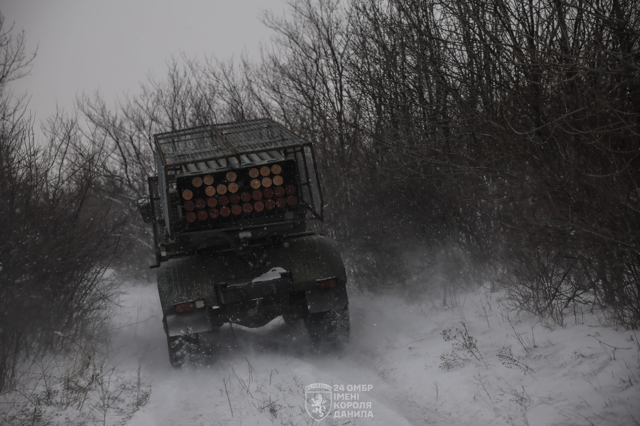 Реактивна система залпового вогню рухається засніженою лісовою дорогою, піднімаючи за собою хмару снігу. На задній частині машини видно пакет пускових труб, а навколо — голі зимові дерева під похмурим небом.