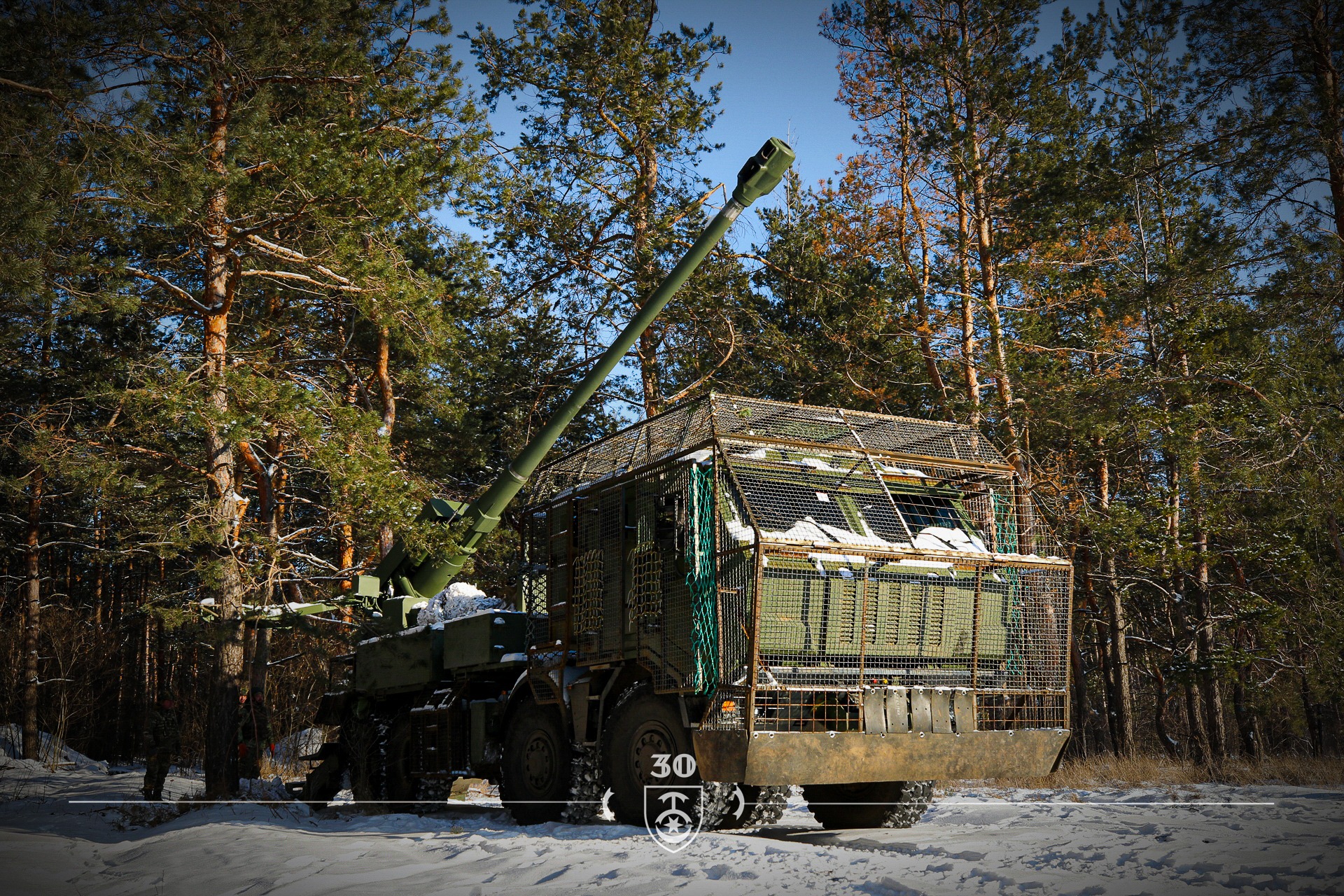 У засніженому сосновому лісі стоїть самохідна артилерійська установка з довгим стволом, спрямованим угору під кутом. Бойова машина обладнана захисною решітчастою конструкцією навколо кабіни та корпусу, а поруч серед дерев видно військових.