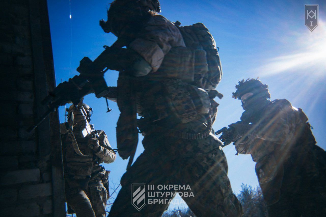 Бійці штурмової групи працюють біля будівлі, прикриваючи один одного та готуючись до входу всередину. Сонячне світло пробивається крізь кадр, підкреслюючи напружений момент бойової роботи.