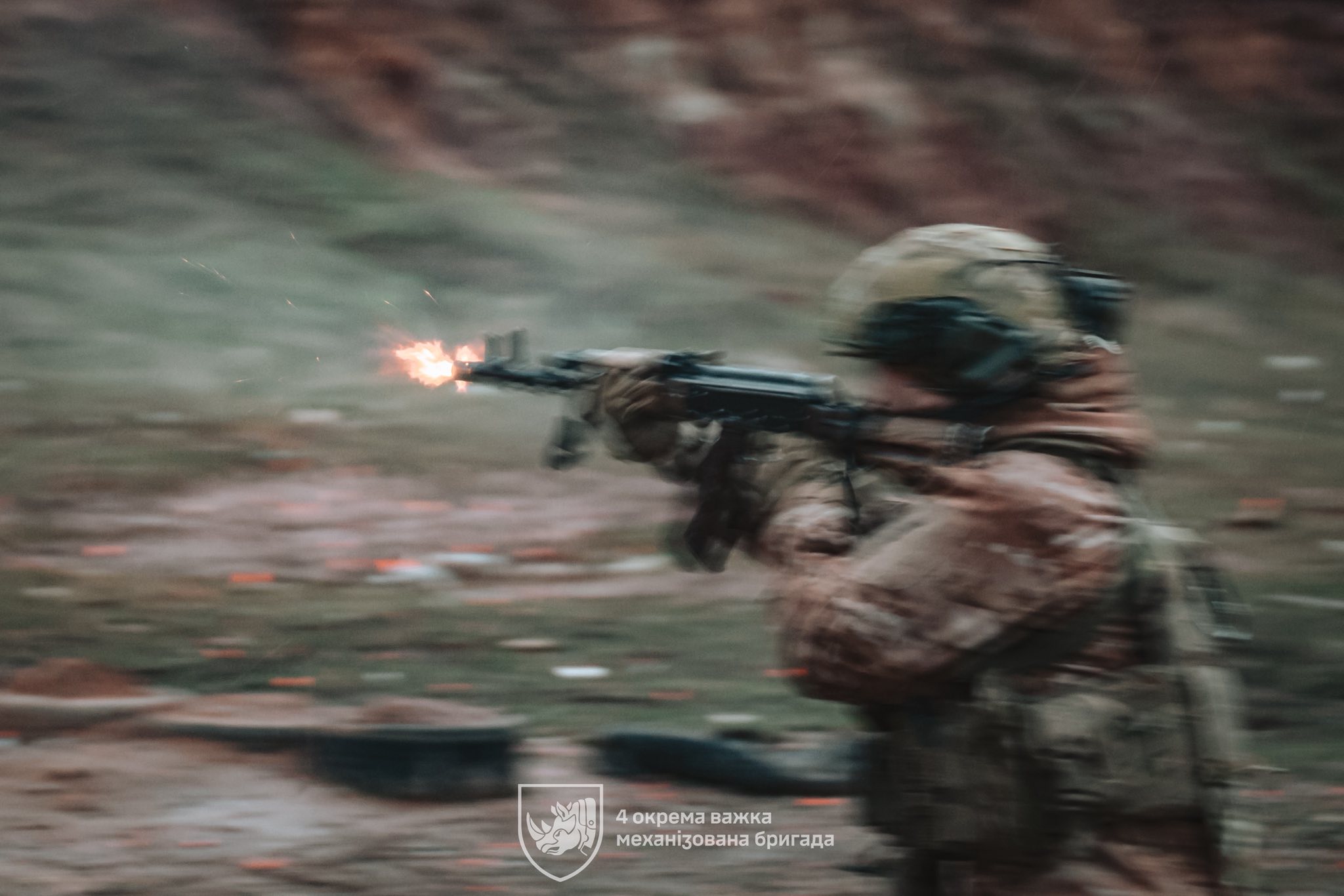 Боєць у захисному спорядженні веде вогонь з автомата під час стрільб, тримаючи зброю на рівні плеча. У момент пострілу біля дульного зрізу видно спалах, а розмитий фон підкреслює динаміку руху та інтенсивність тренування.