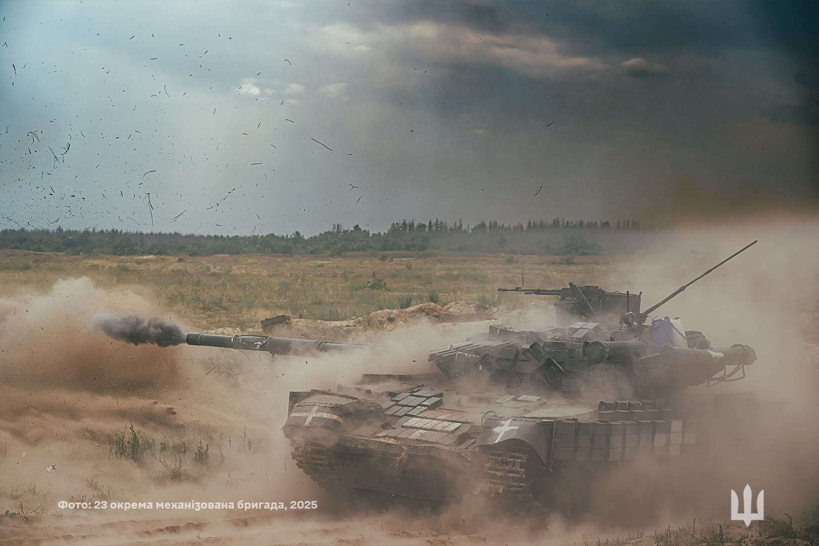 Український танк веде вогонь під час бойових дій на відкритій місцевості, здіймаючи хмару пилу та уламків навколо себе. У момент пострілу зі ствола виривається дим, а над полем розлітаються частинки ґрунту після удару гармати.