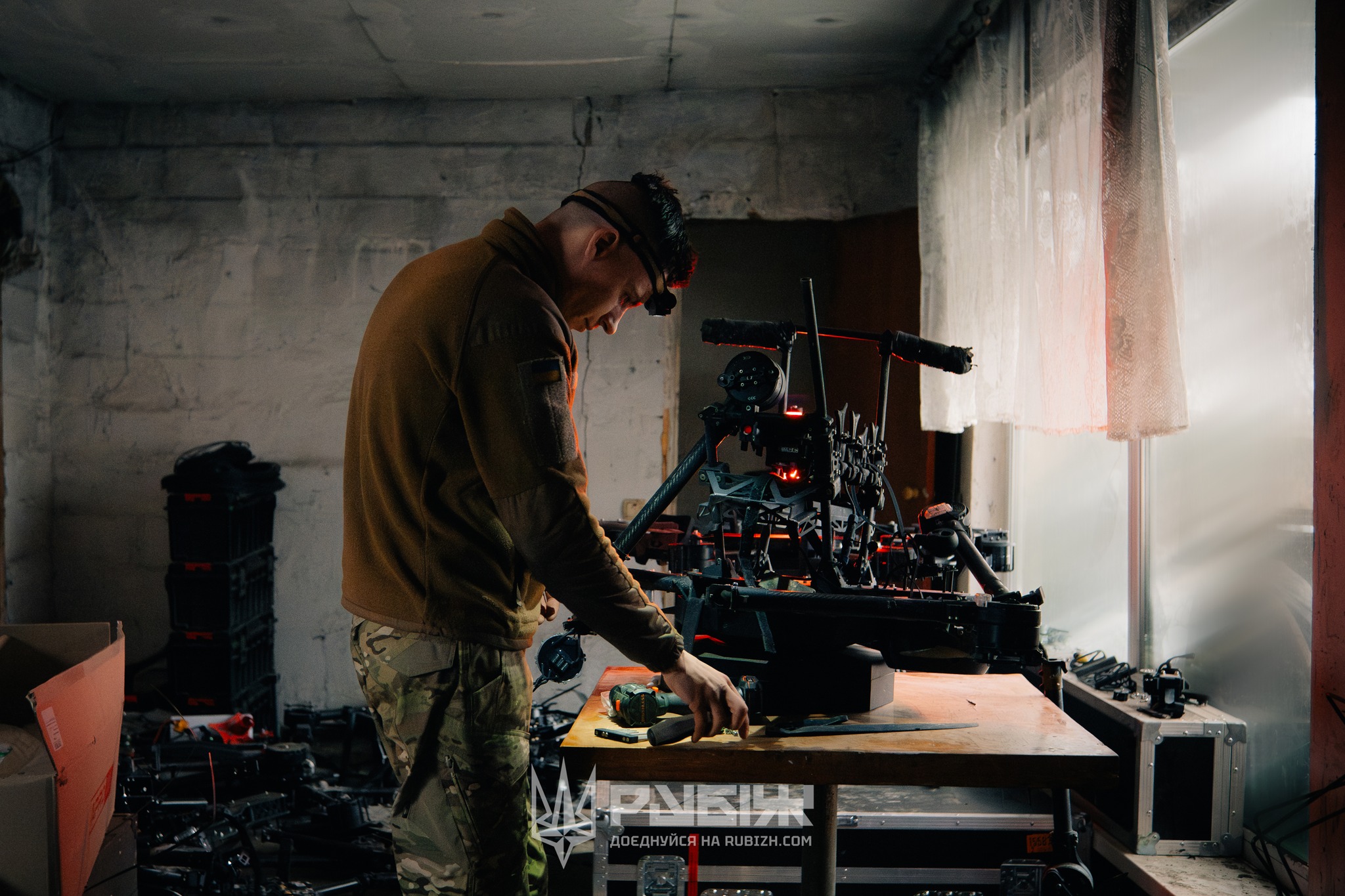 Військовий у приміщенні налаштовує або ремонтує безпілотник, зосереджено працюючи за столом із інструментами. Тьмяне освітлення й технічне обладнання навколо створюють атмосферу польової майстерні.