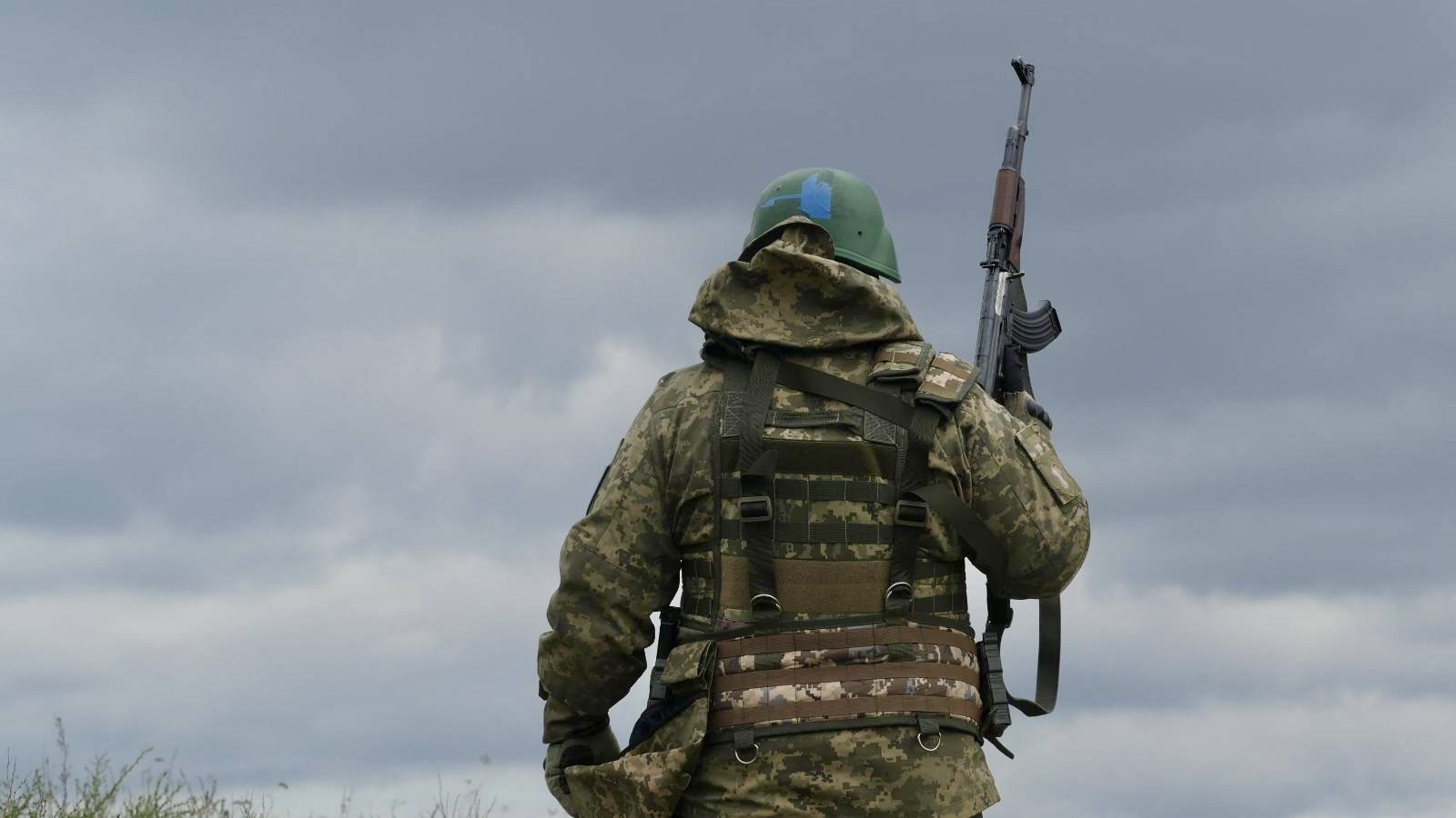 Український військовий у камуфляжі стоїть на відкритій місцевості, тримаючи автомат за плечем. Боєць уважно вдивляється вперед у напрямку поля під похмурим небом.