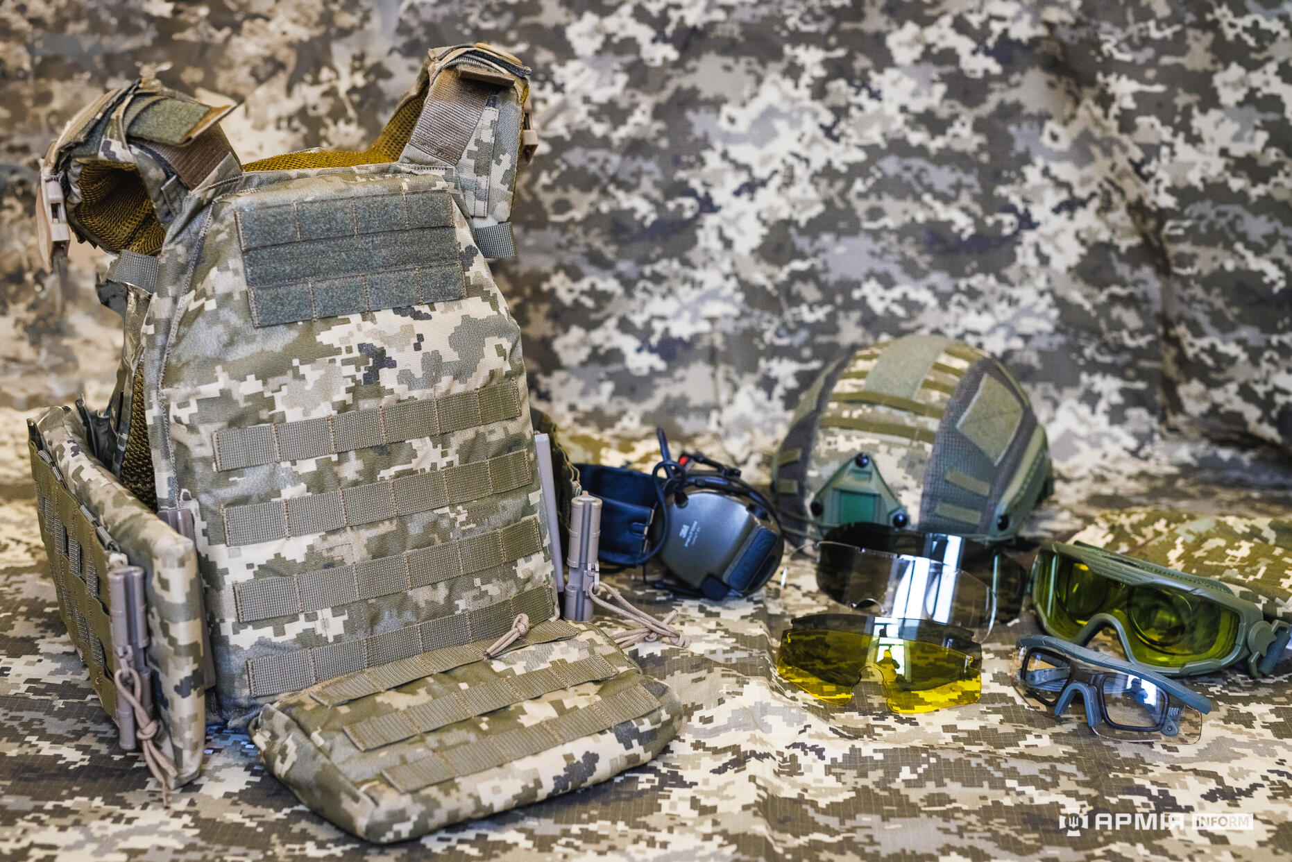 Бронежилет розміщений поряд із шоломом, захисними окулярами та активними навушниками на камуфляжному фоні. Комплект виглядає як повний набір індивідуального захисту військовослужбовця.