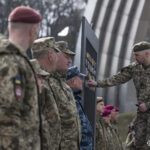 Один із військових прикріплює шеврон до стенда, поки інші уважно спостерігають. Дія відбувається під час церемонії, що має символічний характер.