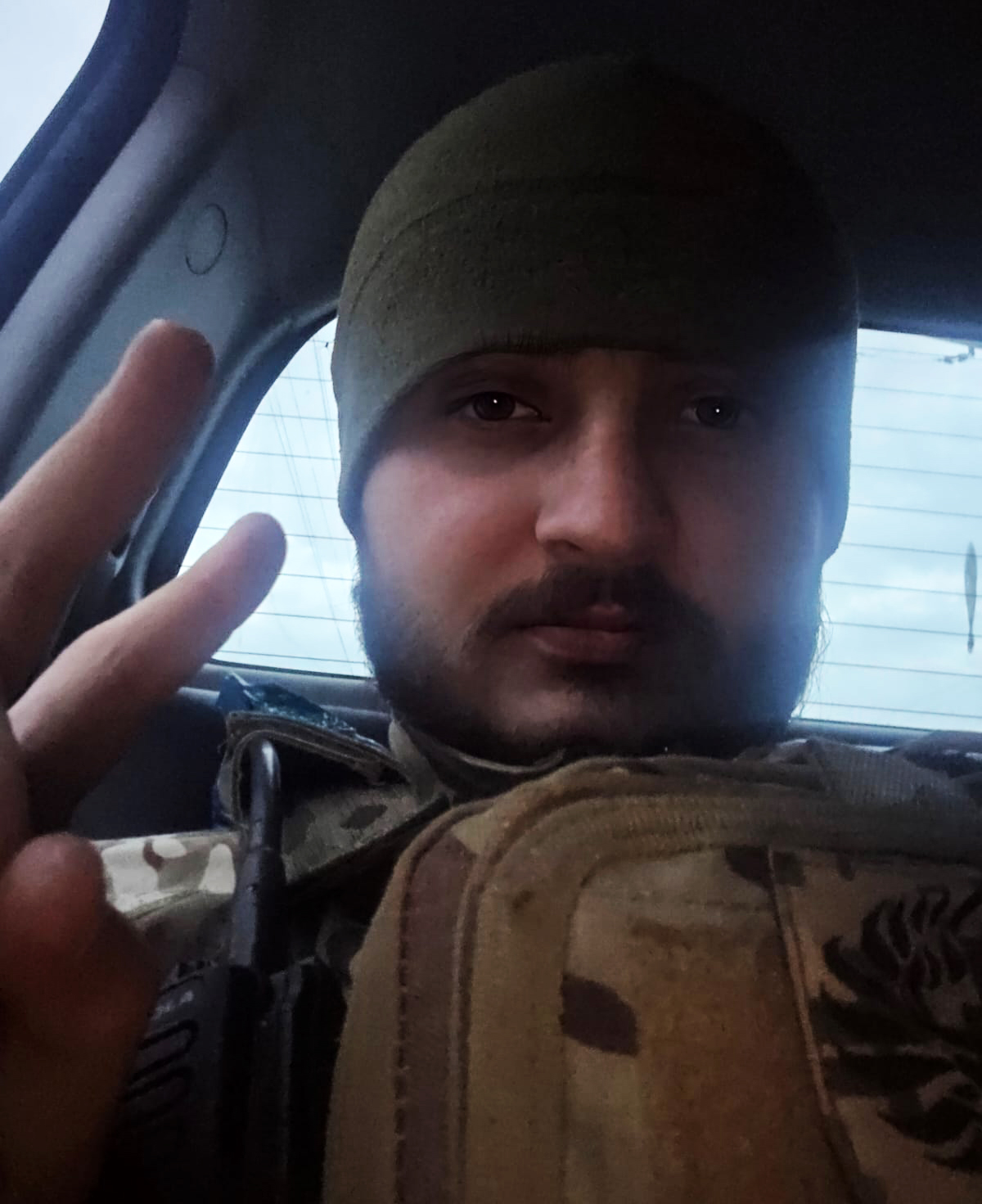 Чоловік у військовій формі робить селфі в автомобілі, піднявши руку з розчепіреними пальцями. На ньому шапка та бронежилет, обличчя освітлене м’яким денним світлом.