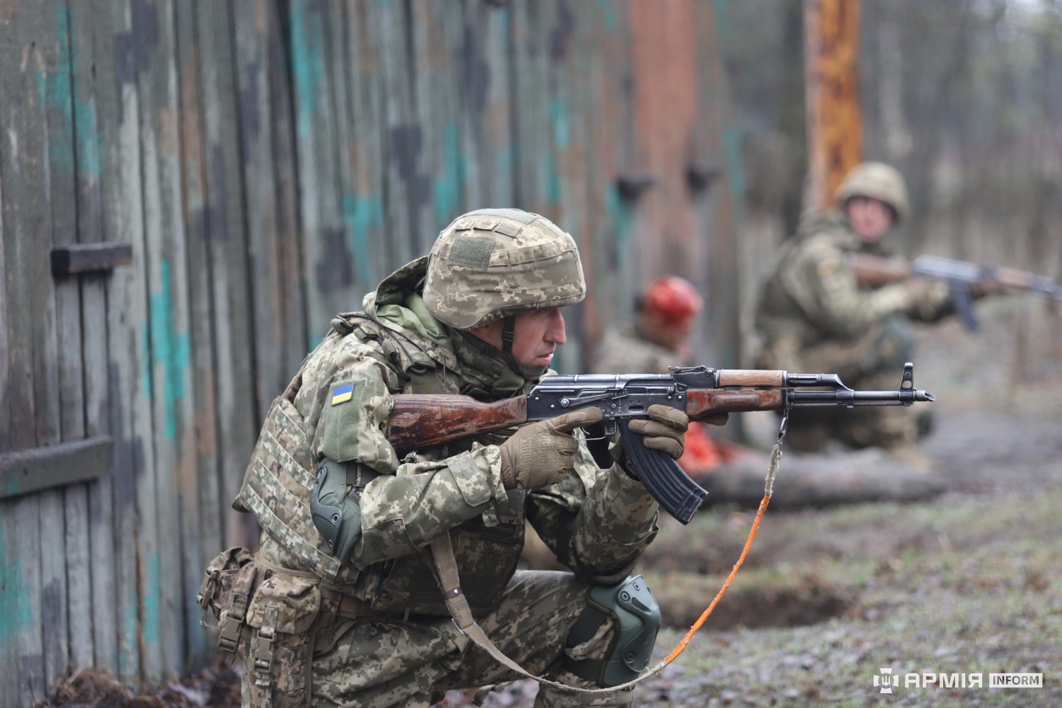 Боєць у камуфляжі, присівши на коліно, веде прицільний вогонь із автомата вздовж укриттів. На задньому плані інші військові займають позиції, контролюючи сектор.