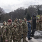 Група військових вишикувалася перед стендом, де вже розміщено десятки шевронів. Поруч працює фотограф, фіксуючи момент події.